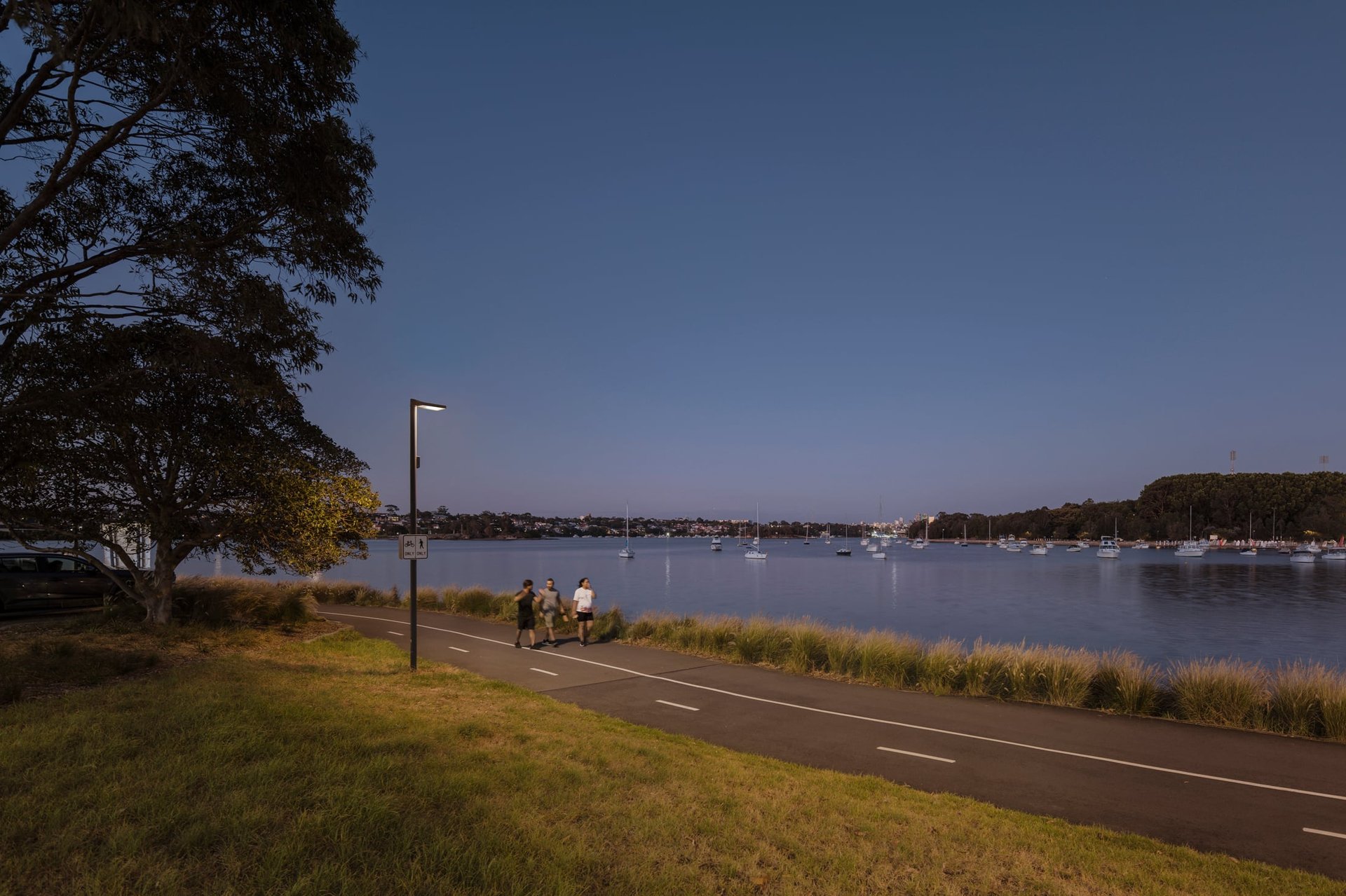 Group walking along the Bay Run path at dusk, enjoying a calm and safe atmosphere enhanced by warm WE-EF street lighting. The smart control system adjusts lighting based on human activity, ensuring efficient, responsive illumination for pedestrians and cyclists.