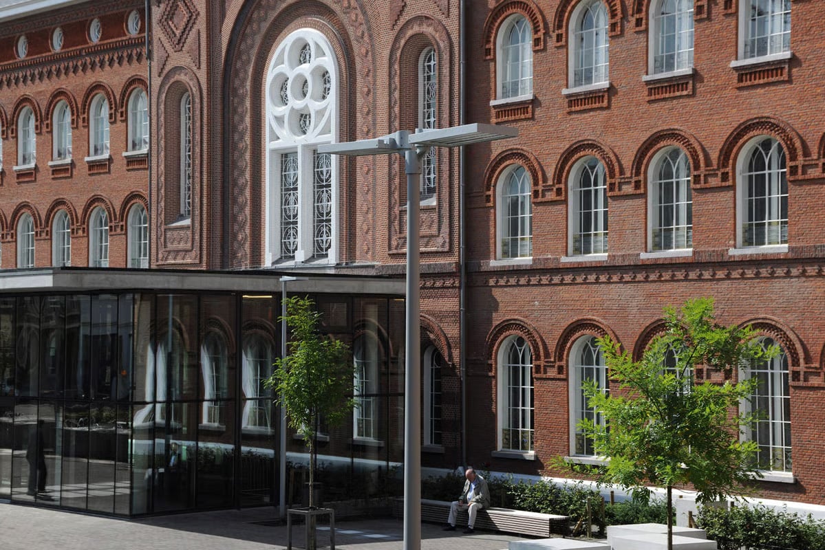 Close-up view of a VFL540 LED luminaire mounted on a slender pole in front of the glass entrance canopy and ornate red brick facade of St. Vincentius Hospital Antwerp, with a person seated on a bench nearby