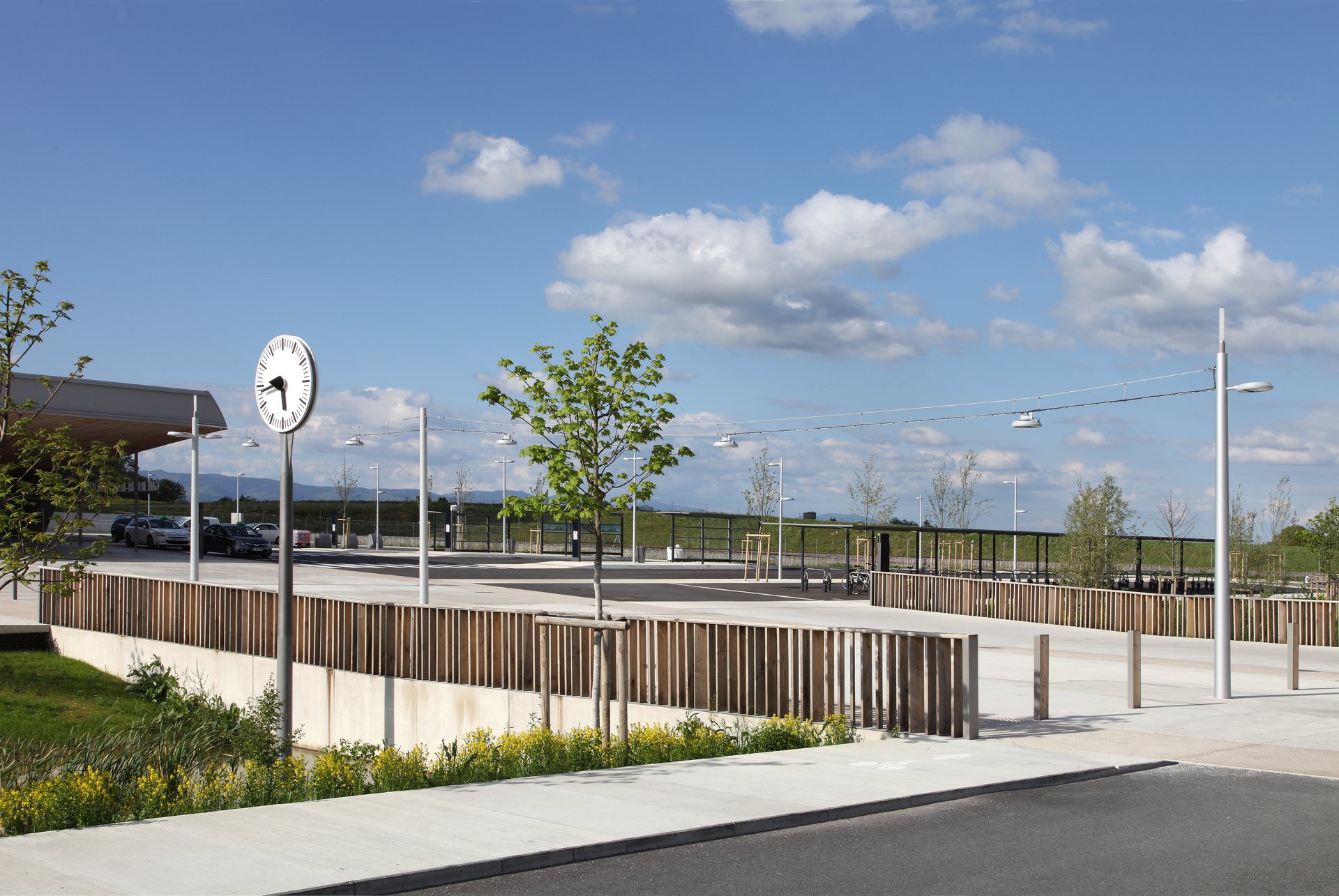 Daytime view of the outdoor area at Belfort-Montbéliard TGV station with station clock, WE-EF RFL LED catenary luminaires, timber elements and views across the surrounding landscape