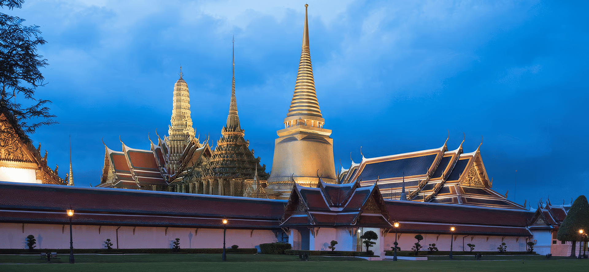 Le complexe du temple Wat Phra Kaew à Bangkok, avec son stupa doré et ses flèches illuminés au crépuscule derrière le mur du palais