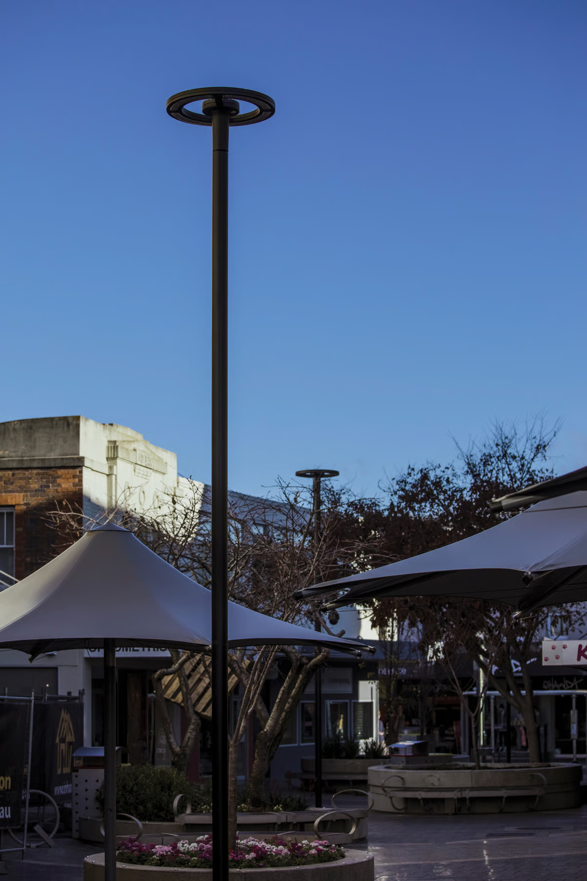 WE-EF CFT540 LED pole-mounted luminaire with minimalist circular design photographed at dusk in Quadrant Mall, showing clean contemporary aesthetic that complements circular planters and white event umbrellas, demonstrating energy-efficient lighting solution with motion-sensor dimming capability for Launceston's City Heart Project