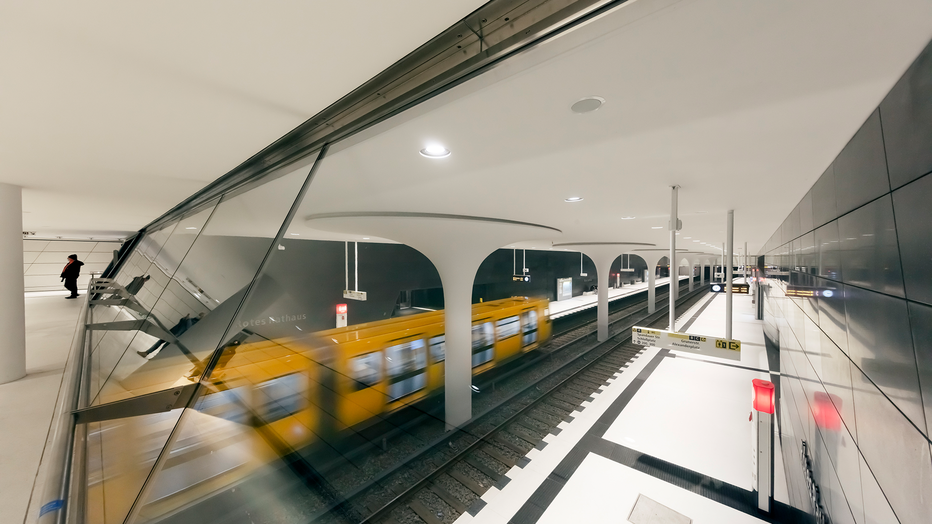 Rotes Rathaus U-Bahn station showing motion-blurred yellow BVG train between dramatic 7x9-meter flared columns in 140-meter prestigious station celebrating Berlin government seat, comprehensive WE-EF LED lighting solution with 70 DOC240 and 100+ DOC220 recessed ceiling luminaires providing symmetrical medium-beam distribution in warm 3000K, ETC140 inground luminaires with DIN 51130 non-slip coating marking platform ends, complex cut-and-cover top-down construction requiring custom housings delivered 2013 years before LED installation 2017, designed by Collignon Architektur with lighting by Licht-Kunst-Licht for December 2020 U5 extension opening