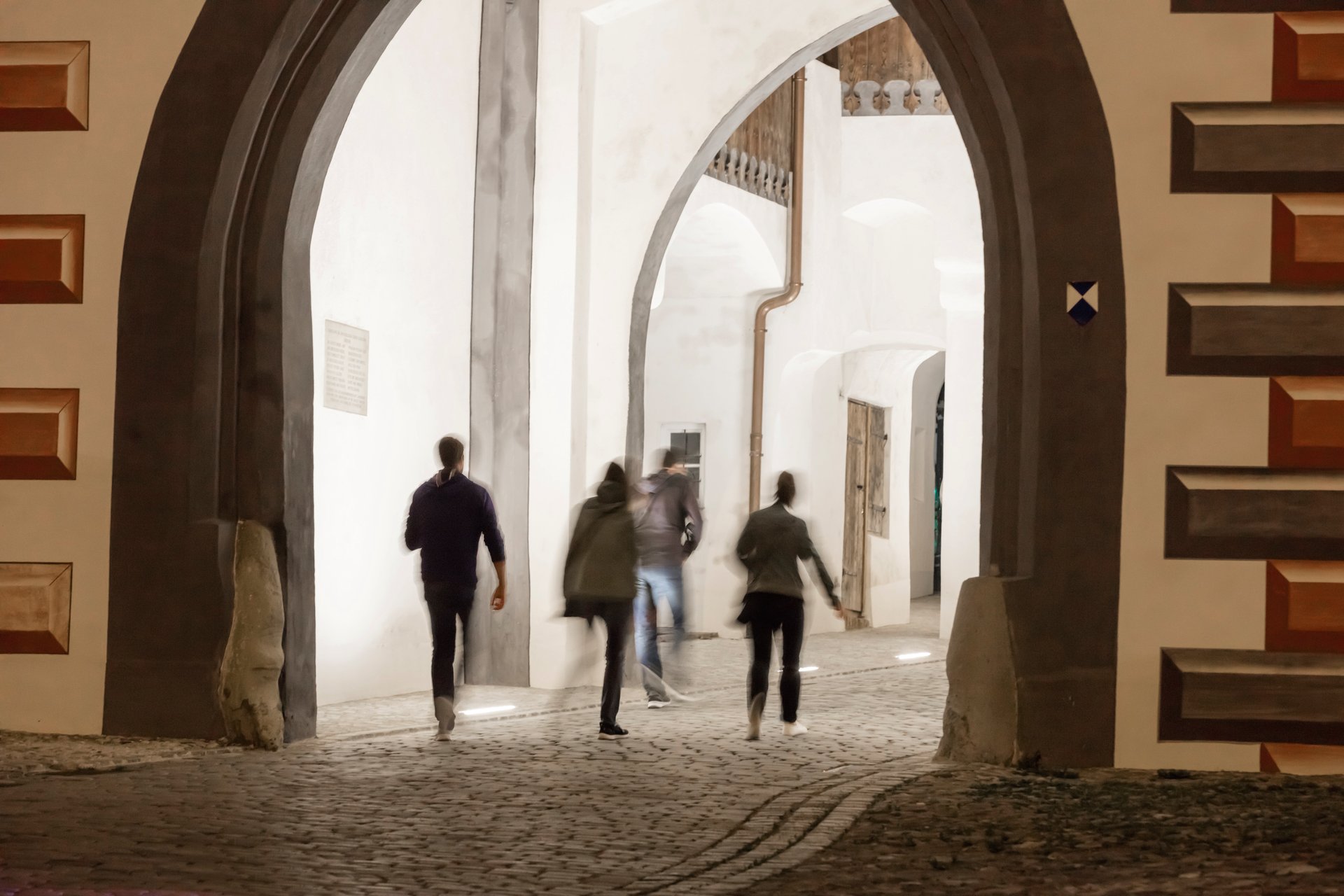 Motion-blurred pedestrians walking through illuminated Gothic archways at Bayertor at night, with WE-EF linear inground lighting providing safe pathway guidance while dramatically highlighting the historic vaulted architecture and ornate wooden doors