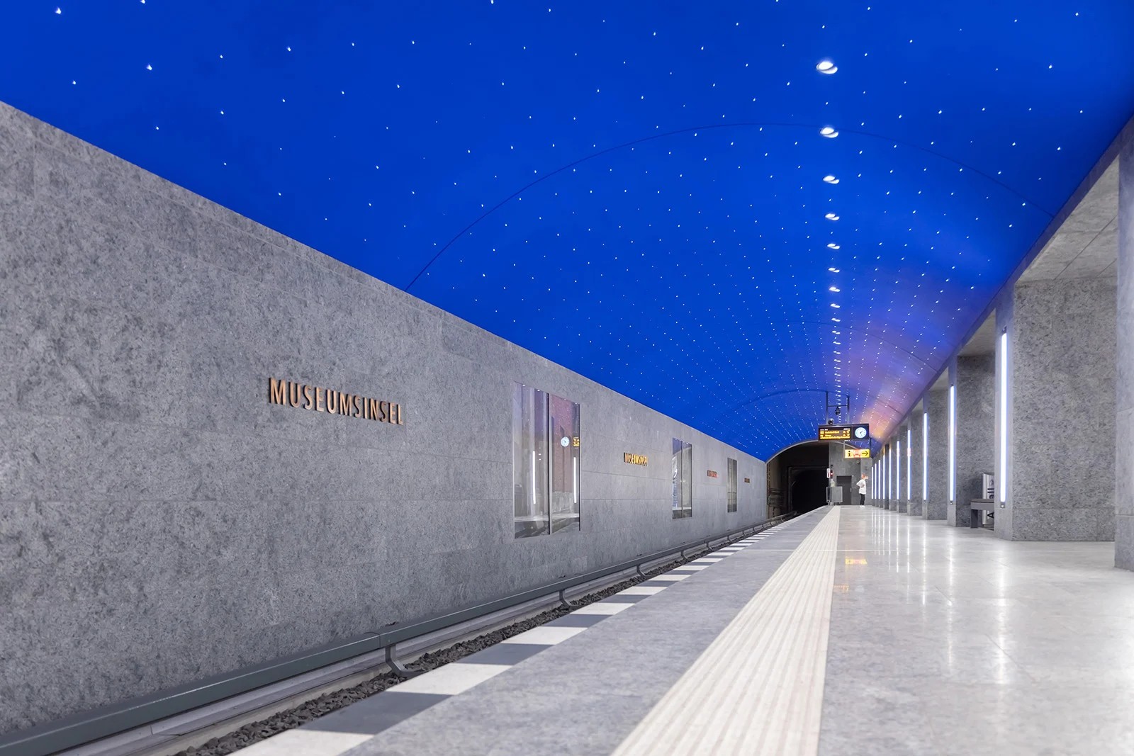 Contemporary entrance to Museumsinsel U5 station featuring minimalist granite parapet design with integrated WE-EF VLR100 series linear luminaires providing functional downward wall wash lighting into entrance void, transparent glass-enclosed escalator descent visible beneath iconic Berlin U-Bahn signage, architectural lighting solution by Thomas Spreen/High Light supporting Max Dudler's design concept for 2020-completed station serving Museum Island's cultural heart beneath Spree Canal