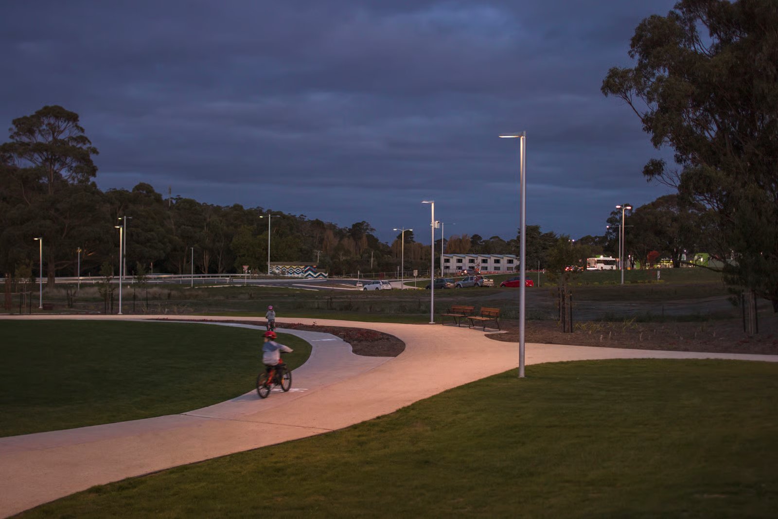 Kingston Parklands playground and pathways at twilight with children on bicycles, WE-EF VFL530 pole-mounted LED luminaires with P65 and S65 optics providing safe pathway illumination in warm 2700K, precise light distribution ensuring visitor safety and visual comfort while minimizing light pollution impact on surrounding native eucalyptus bushland habitat designed by Playstreet landscape architects on former Kingston High School site