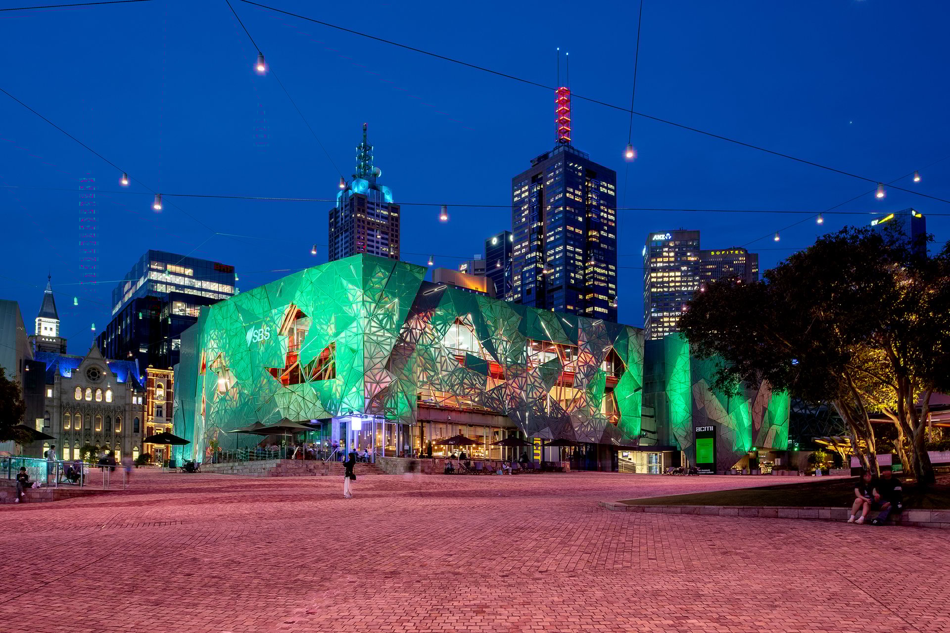 Federation Square's main plaza at blue hour with iconic geometric buildings illuminated in vibrant emerald green using WE-EF FLC200 RGBW color-changing floodlights, Melbourne's CBD skyline featuring illuminated towers in background, catenary lighting overhead, historic cathedral visible left, brick paving across public space, demonstrating the $20M Victorian Government lighting upgrade designed by ARUP for enhanced evening visitor experience