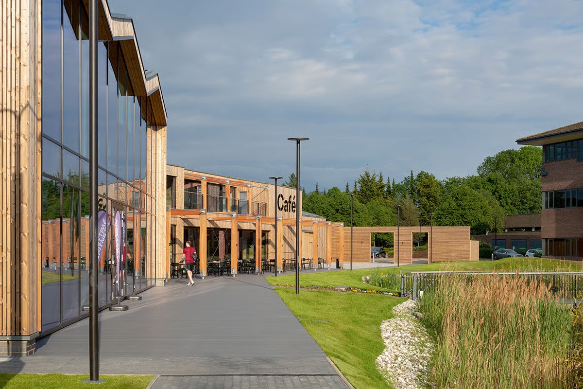 Croxley Park contemporary office building showing timber-clad facade with comprehensive WE-EF CFT540 lighting along pedestrian pathway, clear modern design language of ring-shaped pole luminaires harmonizing with campus architecture designed by ESA London, durable energy-efficient LED technology with warm white 3000K supporting operator's sustainability goals, parkland setting near Watford with good transport links creating attractive working conditions where employees can enjoy café, gym, day-care and other amenities