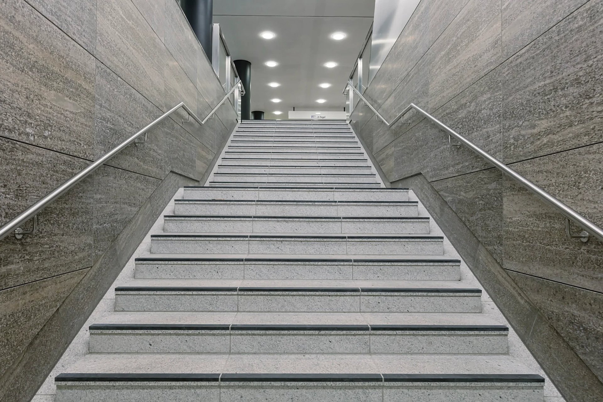 Wide stairway at Unter den Linden station showing timeless exposed concrete and shell limestone architecture, 410 modified WE-EF DOC240 LED luminaires with project-specific mounting elements optimized for installation cavities created during cut-and-cover construction, custom acrylic glass covers bolted and wire-cable-secured against vibration providing uniform 3000K warm white light with characteristic soft rim illumination, designed by High Light Berlin for prestigious 2020-completed U5 line extension serving German capital
