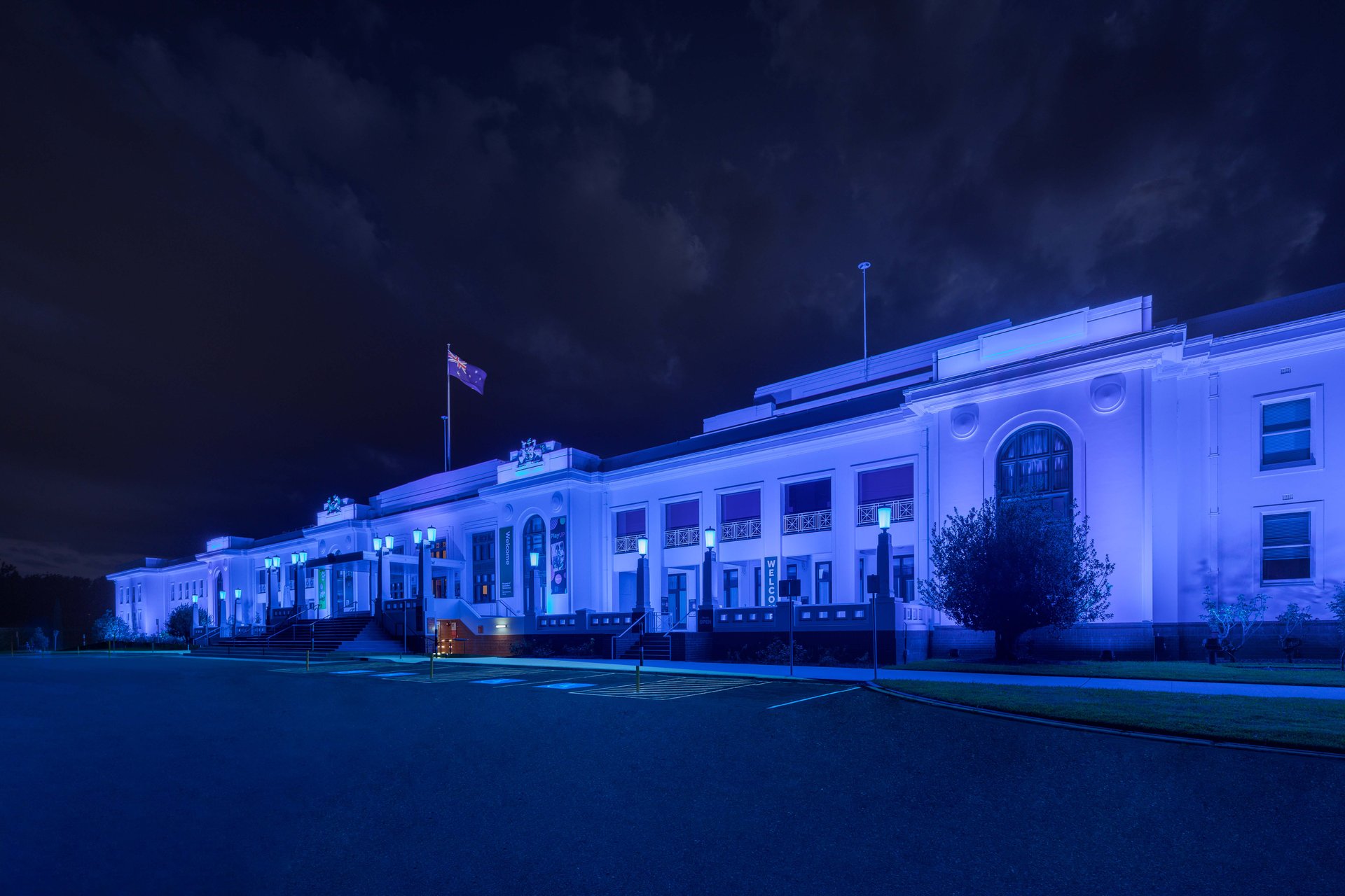 Old Parliament House Canberra front facade in deep blue lighting showing WE-EF color-changing luminaires providing uniform wallwashing across entire width of provisional parliament building designed to serve 50 years but actually hosting Federal Parliament from 1927-1988, combination of FLC200-CC projectors and VLR100-CC surface-mounted linear wallwashers suitable for outdoor architectural facade accentuation, DMX integration enabling programmed lighting scenarios for Museum of Australian Democracy