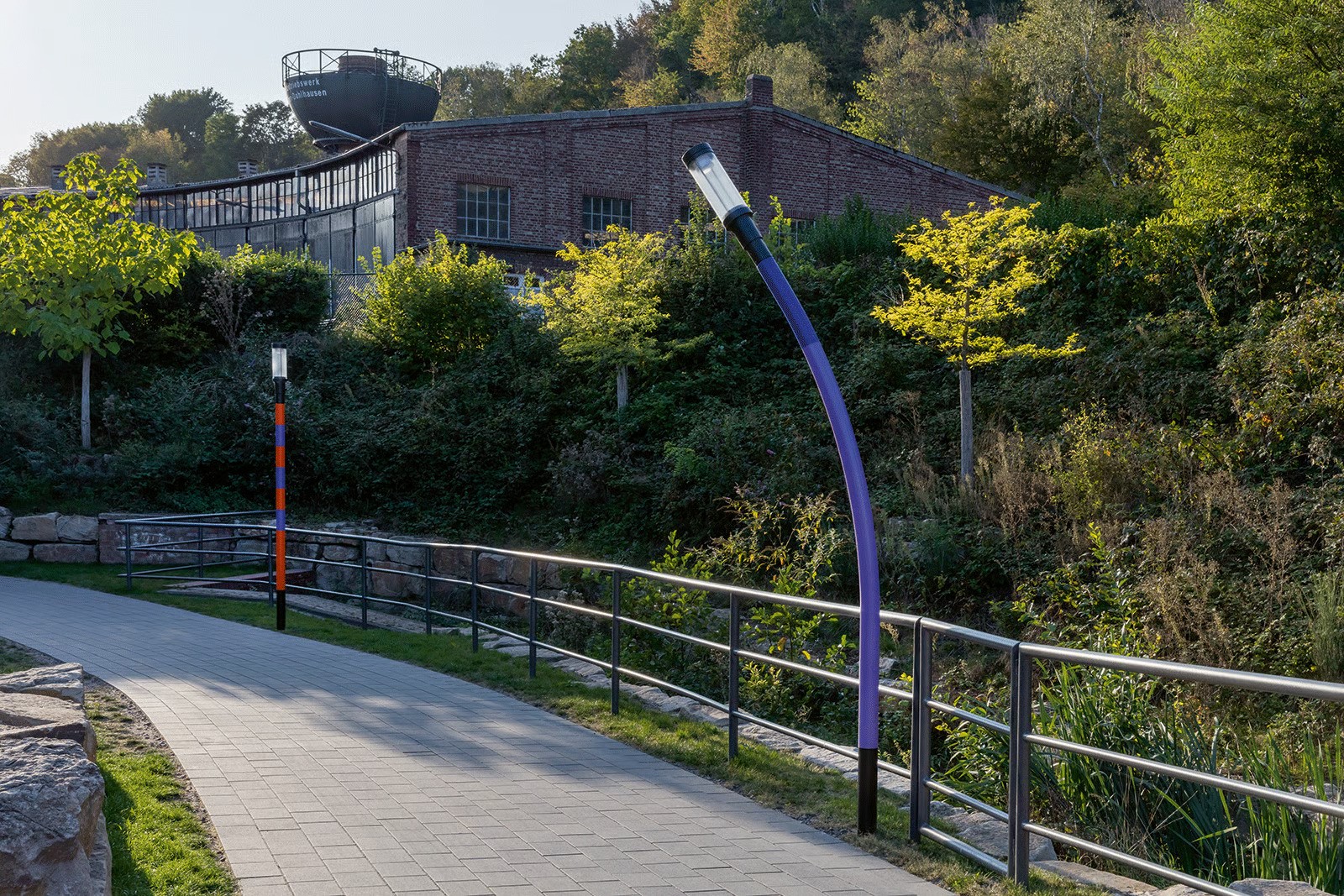 Barrier-free ramp at Bochum Dahlhausen tunnel entrance featuring customized WE-EF ZFT440 LED street luminaires with individually colored poles (purple and orange-blue visible) bent in unique postures, each of 18 luminaires providing symmetric C60 beam distribution in 4000K with functional reliable lighting while quirky design brings light and humor to harsh industrial environment, former railway depot buildings visible above, demonstrating WE-EF expertise and flexibility in producing custom outdoor luminaires for 2020 renovation transforming century-old scary underpass into safe welcoming thoroughfare