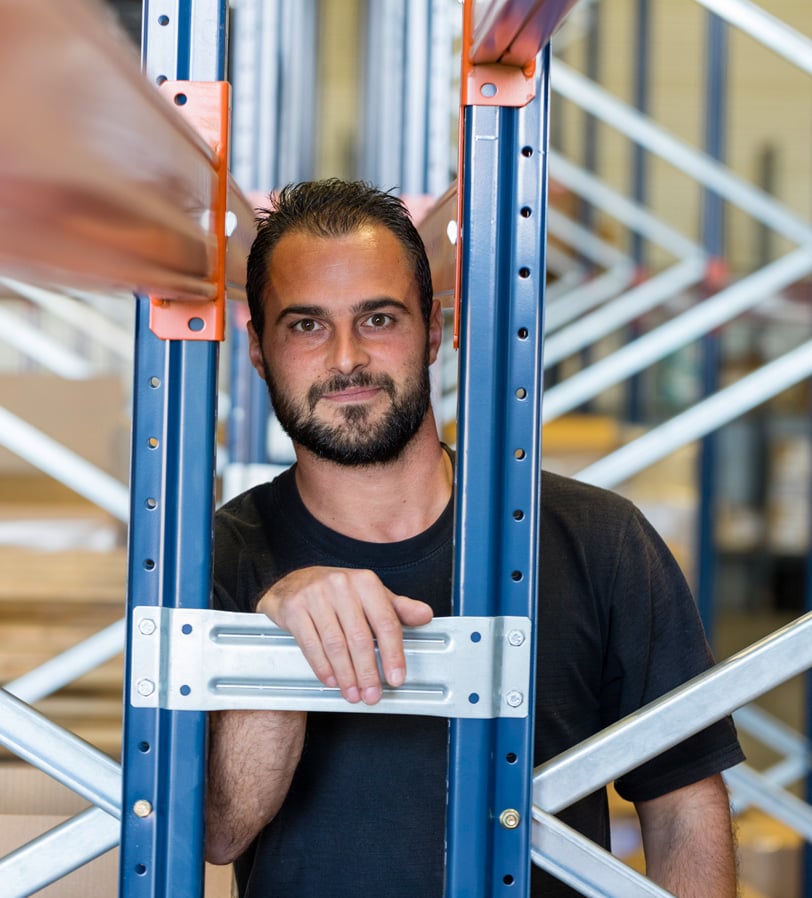 Collaborateur WE-EF dans l'entrepôt, appuyé contre un rayonnage à palettes bleu et orange, regardant l'objectif