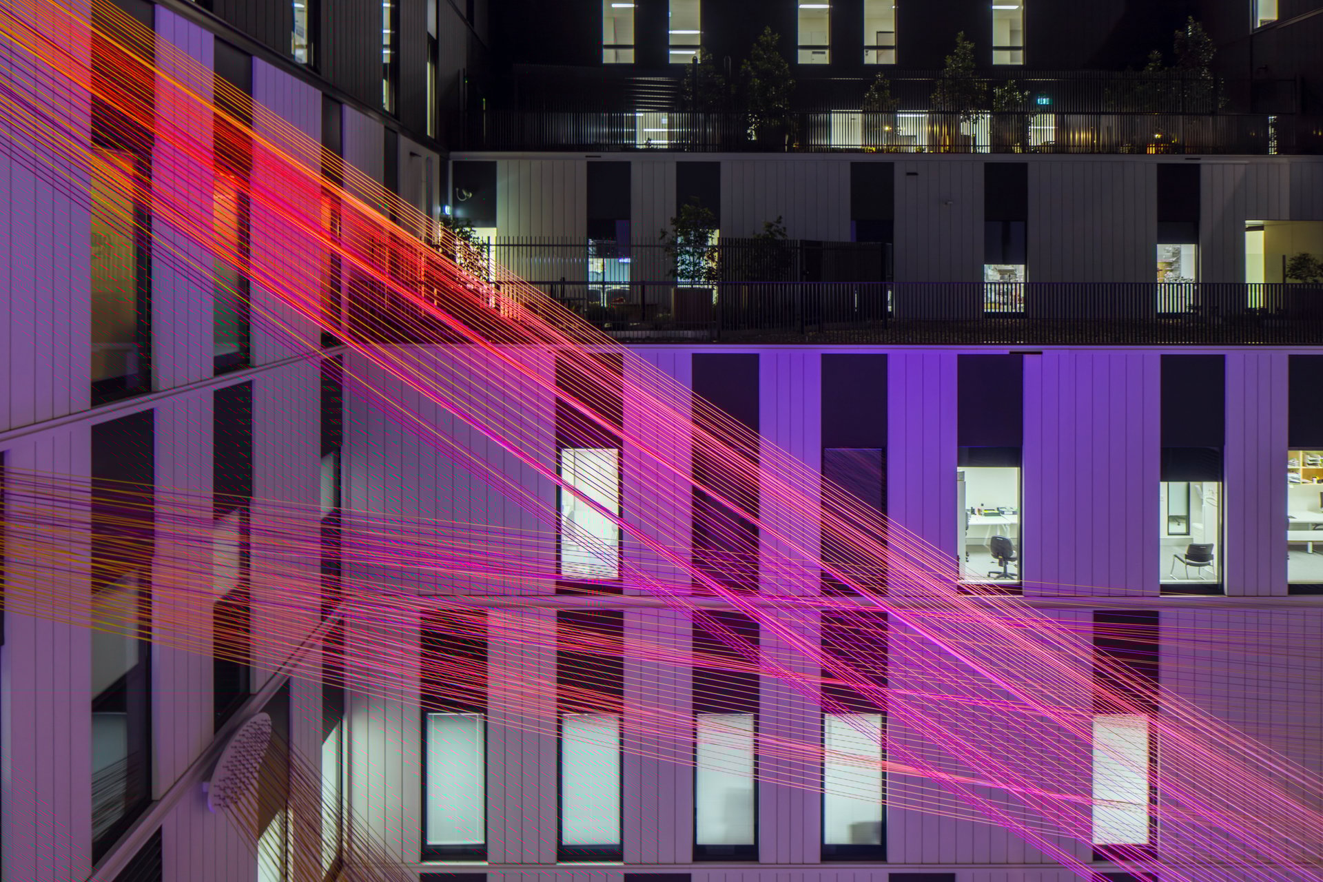 Leuchtende, pink‑ und rotfarbene Lichtstränge spannen sich nachts zwischen den Fassaden eines modernen Gebäudekomplexes. Die farbigen Linien erzeugen ein schwebendes, dynamisches Muster vor den hellen Fensterflächen und den vertikalen Paneelen der Architektur.