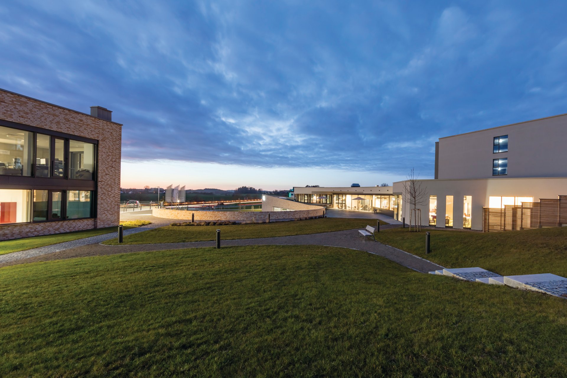 das flax Hotel campus at blue hour showing multiple buildings with light-colored clinker brick facades designed by FG-Architektur in campus-like topography nestled in Allgäu Alpine foothills, comprehensive WE-EF lighting including KTY234 bollards for pathways, VFL530 dual-arm pole luminaires for driveway, and ETV130 linear inground wallwashers highlighting facades, creating urban island feel in rural German setting near Dietmannsried