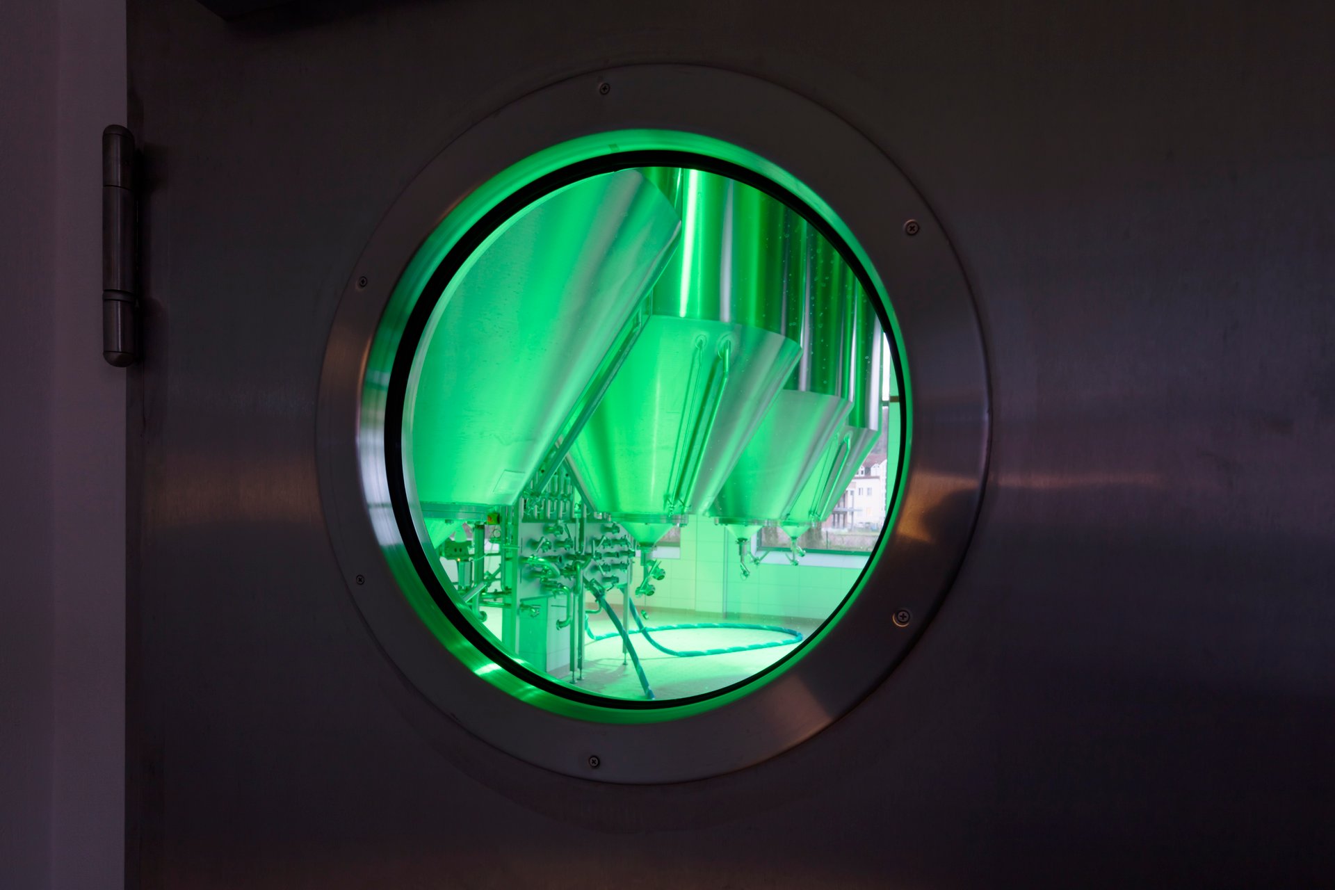 View through stainless-steel circular porthole into Schattenhofer Brewery fermentation tank room illuminated in bright green by WE-EF FLC220-CC RGBW projectors, gleaming stainless-steel fermentation tanks and piping visible, demonstrating transparency and quality awareness emphasized by large windows and magical colored light showcasing modern brewing technology in partnership with nearby Gutmann brewery from Titting