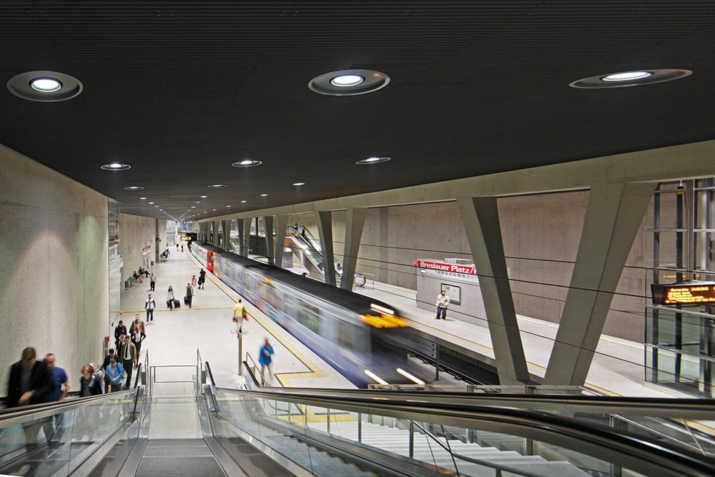 Belebter Bahnsteig des U-Bahnhofs Breslauer Platz in Köln mit einfahrender Stadtbahn, WE-EF Deckeneinbauleuchten, Rolltreppen und Fahrgästen
