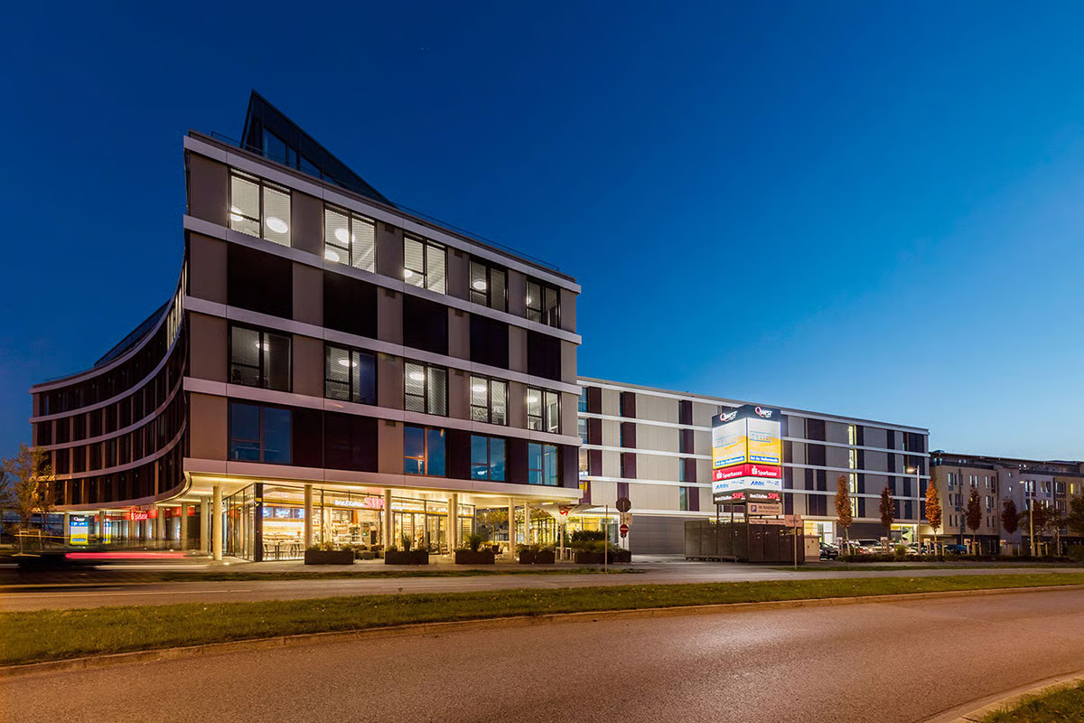 Q.West Ingolstadt mixed-use development at blue hour showing curved modern architecture with illuminated retail spaces at ground level, residential units above, and WE-EF RFL530-SE street lighting along roadway