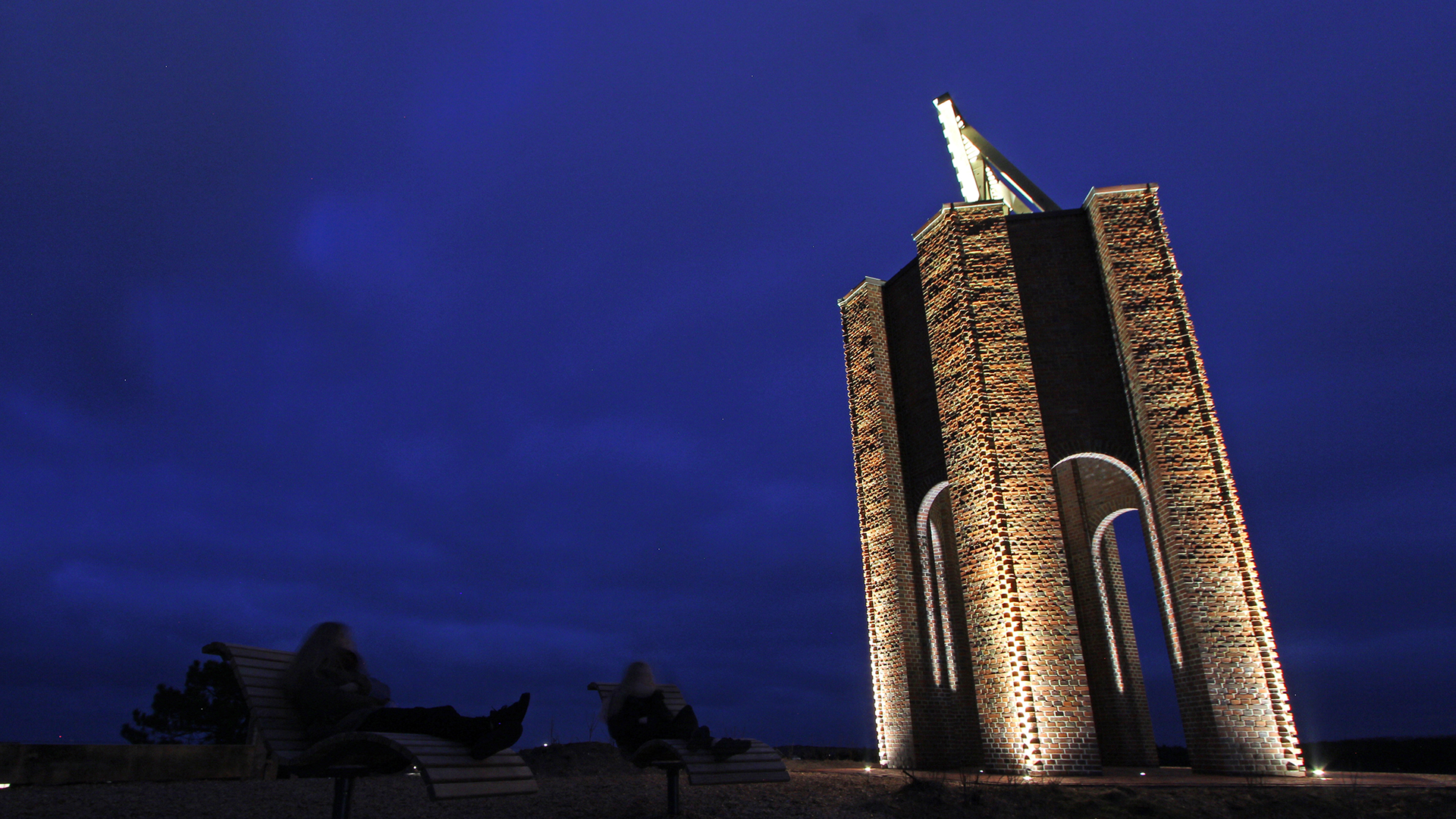 Cape Norderney beacon at night showing historic 12-meter hexagonal brick structure erected 1871 atop 15-meter dune with dramatic high-contrast lighting by Oliver Christen OC-Lichtplanung, six angular columns illuminated by precisely aligned WE-EF ETC120-GB gimbal-mounted inground luminaires with symmetric very-narrow-beam optics accentuating lively brickwork texture and round archways, triangular wooden slat sea marker floating above structure, island coat of arms symbol since 1928 transformed from functional 1848 seafarer safety beacon to illuminated tourist landmark in East Frisian Wadden Sea