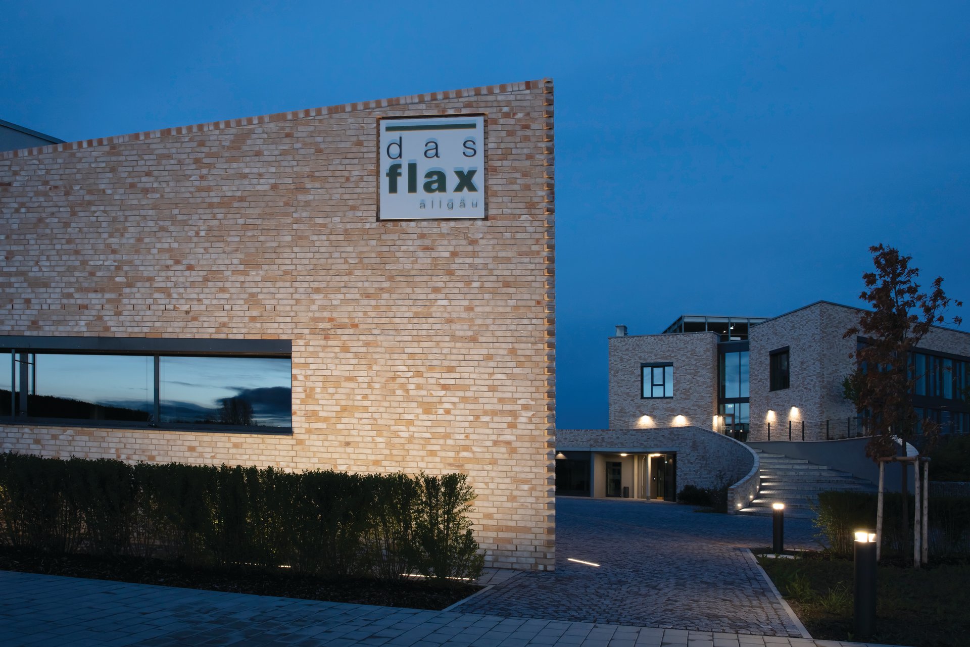 das flax Hotel entrance facade at twilight showing light-colored clinker brick wall with illuminated hotel logo, uniformly lit by WE-EF ETV130 linear LED inground wallwashers providing precise architectural accent lighting, demonstrating streamlined contemporary hotel concept appealing to style-conscious business and leisure travelers in economy segment, completed 2020 by Voralpenresort Besitz GmbH