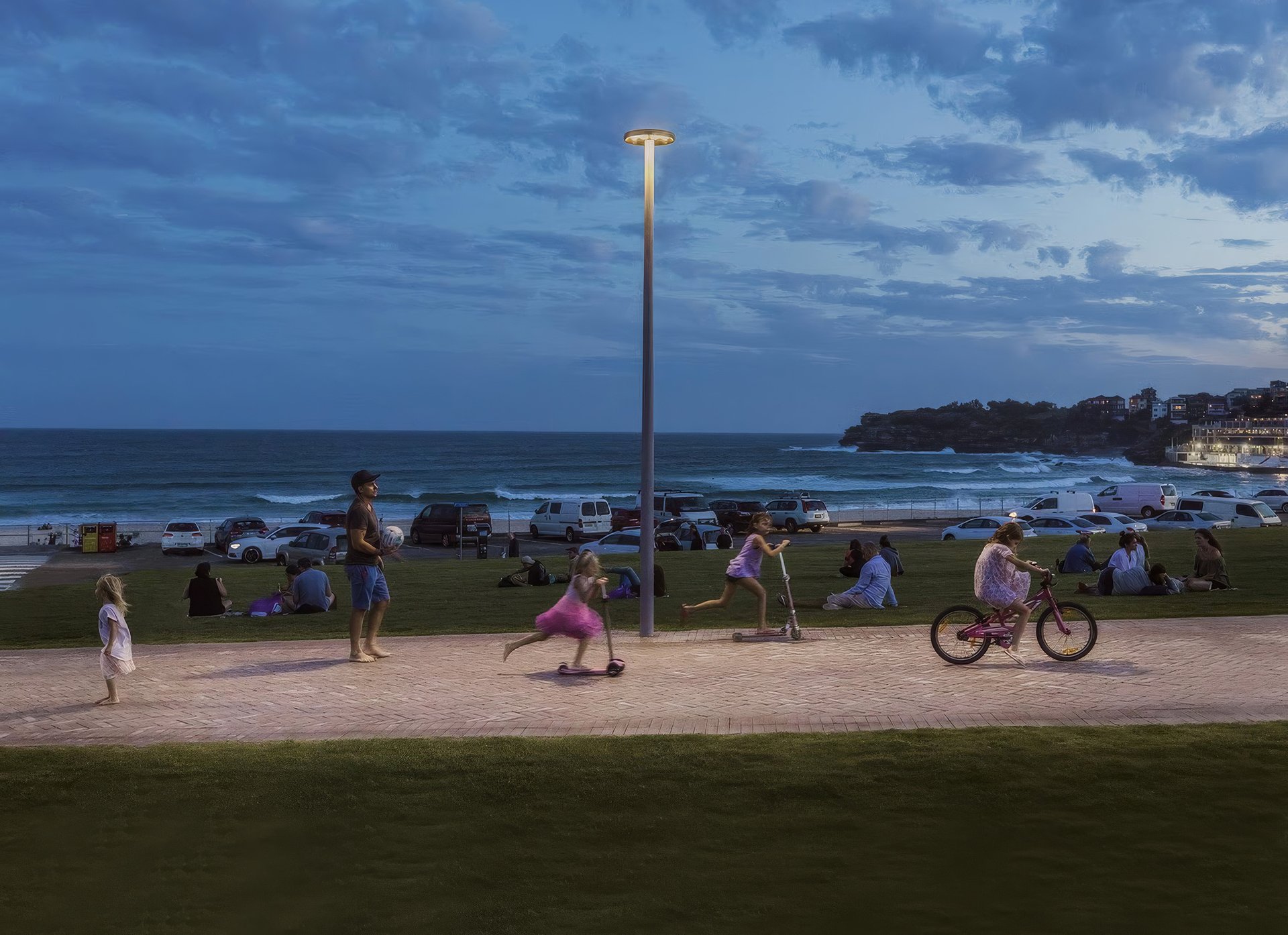 Colonne lumineuse WE-EF éclairant au crépuscule un chemin en bord de mer à Bondi Beach, où des enfants circulent à trottinette et à vélo