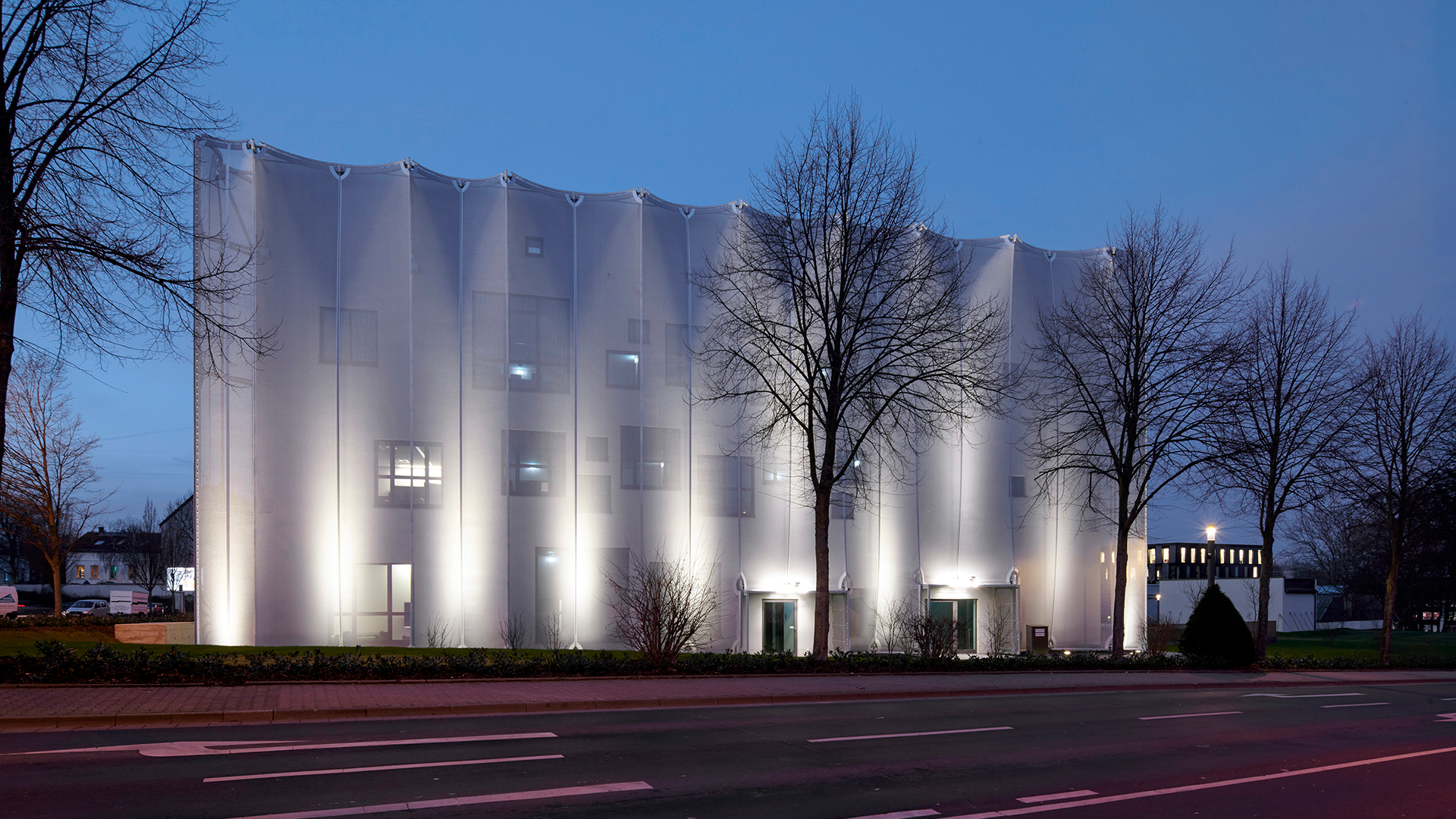 Textile Academy NRW Mönchengladbach at twilight showing comprehensive WE-EF facade lighting using ETC140-GB gimbal-mounted inground uplights and FLC141 projectors with precision very-narrow-beam LED optics and honeycomb louvres for optimum glare control, intensity-adjusted illumination showcasing transparency of 2,100-square-meter textile membrane composed of individual fabric lengths forming irregular wave pattern, lighting design by Maass-Licht revealing complex architectural language of shimmering silver sheath by Slapa Oberholz Pszczulny SOP wrapping three-story private vocational college for textile professions in traditional German textile manufacturing center