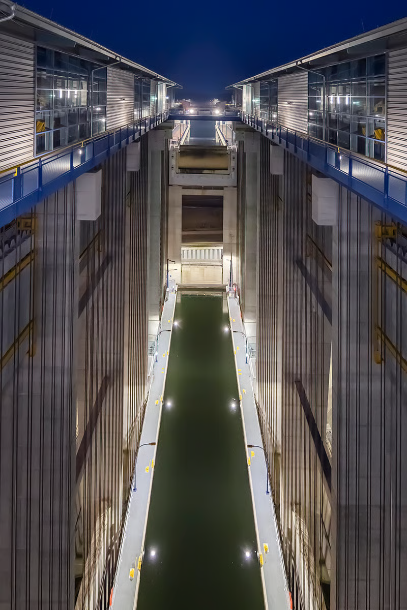 Vertical view into Niederfinow ship lift's 9800-tonne water-filled trough showing WE-EF PFL540 luminaires with customized hybrid optics featuring asymmetric side-throw LED modules (S60 front, S70 rear rotated 180°) mounted on 6-meter poles along trough walls, FLC141 LED projectors with medium-beam characteristics providing additional targeted illumination of critical holding gate areas, all in neutral white 4000K, control station visible above at 50-meter height