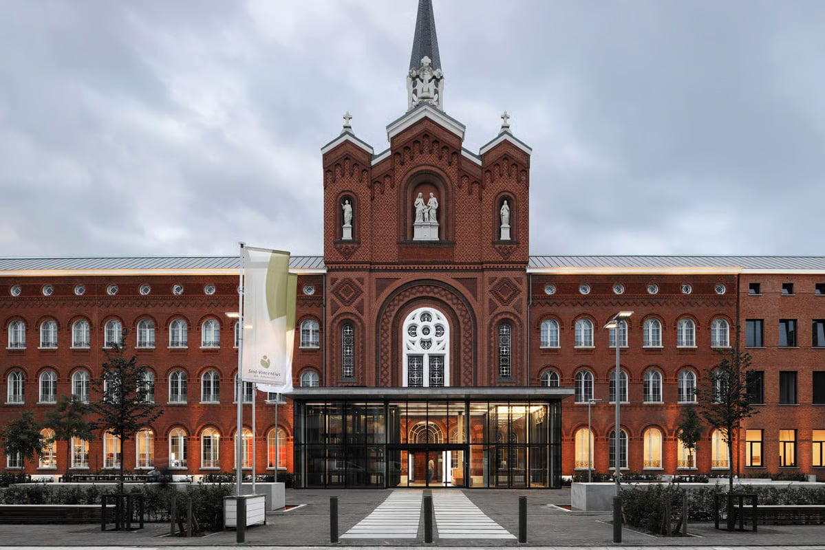 Illuminated facade of Sint-Vincentius Hospital Antwerp at dusk, showing the symmetrical red brick historic building with central Gothic chapel tower, white stone statues, rose window, and a modern glass entrance canopy, with VFL540 LED pole luminaires and the hospital's flag in the foreground under an overcast sky