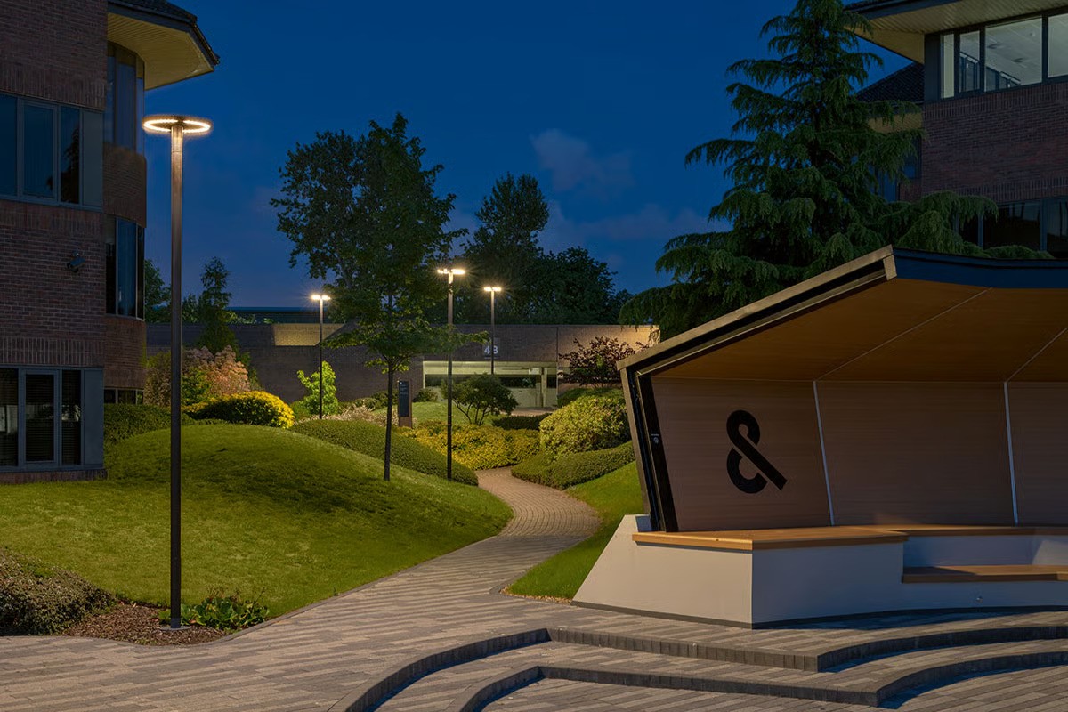 Croxley Park pathway at evening showing WE-EF CFT540 LED pole-mounted luminaires with ring-shaped design using OLC One LED Concept multi-layer lens technology ensuring uniform light distribution with very good visual comfort, RFC technology with UV-stabilized PMMA or PC contour cover following lens shape reducing internal reflection to absolute minimum, warm 3000K illumination creating pleasant after-work environment, contemporary angular wall sculpture visible demonstrating architectural quality determining attractiveness of office park
