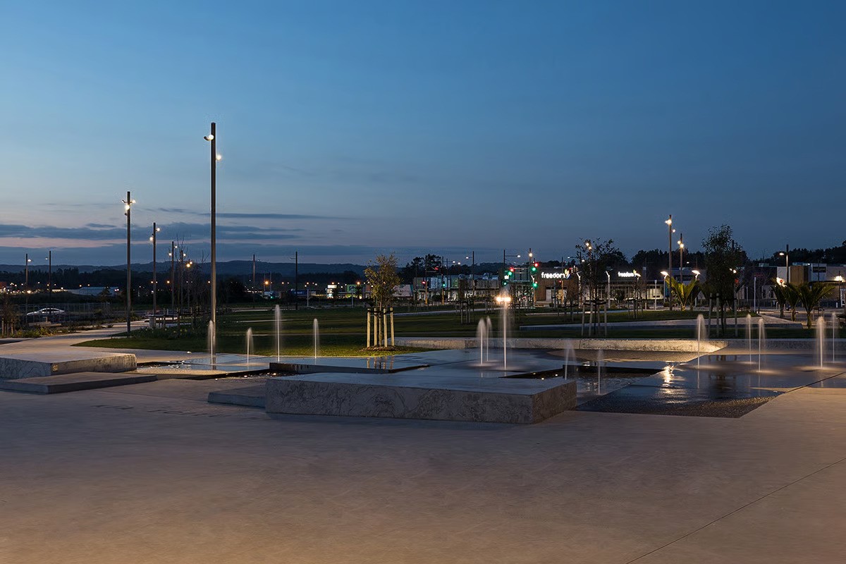 Te Hauāuru Park water feature at evening showing reflective surface with comprehensive WE-EF lighting designed by MHL Auckland, FLC121 projectors with drivers in base of poles accessed via flange door for easier maintenance providing clean uncluttered appearance, lighting scheme accommodating future growth of Westgate town centre masterplan including additional shopping mall level, carpark and office buildings, completed 2017 for Auckland Council creating green space adjacent to award-winning Kopupaka Park in one of Australasia's biggest shopping precincts
