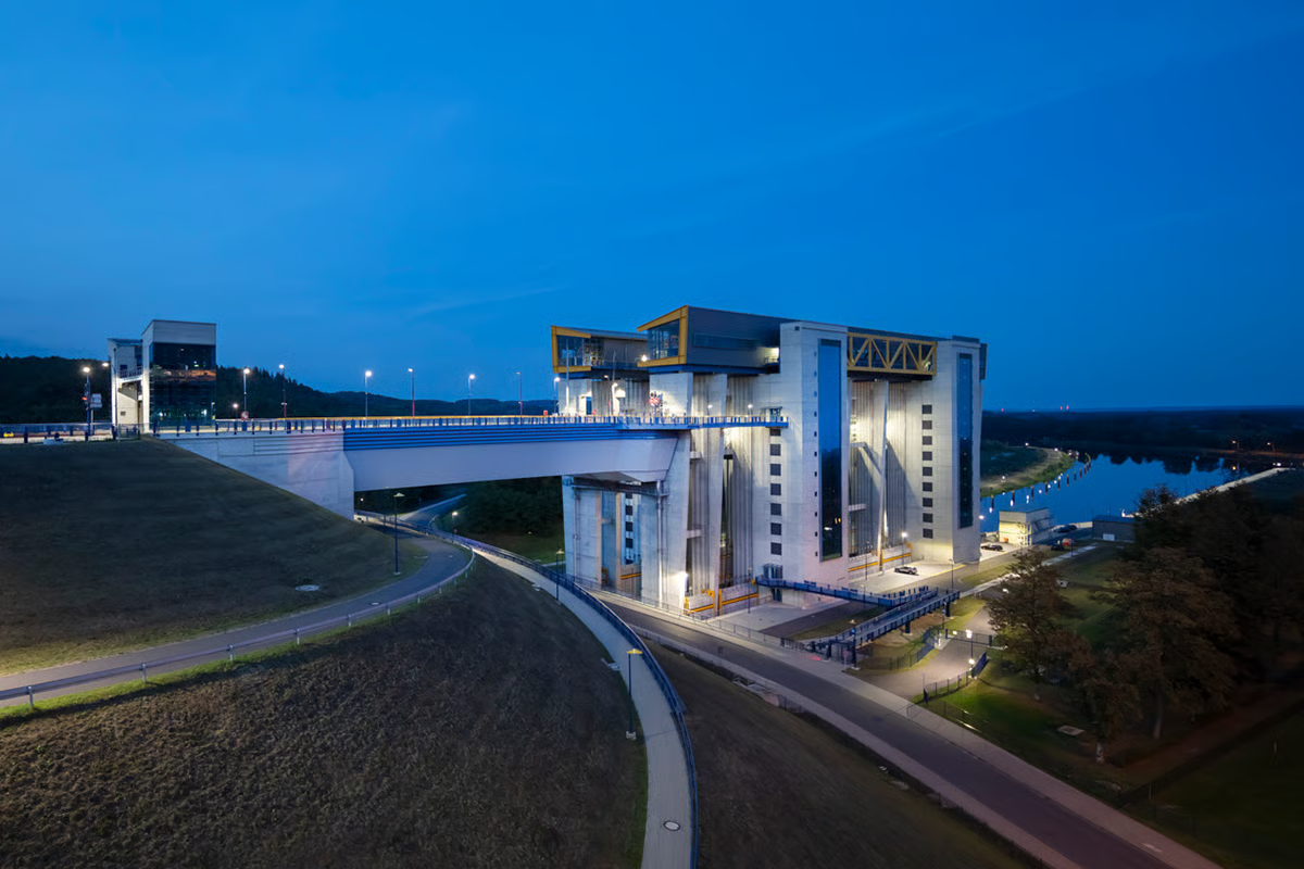 Niederfinow ship lift illuminated at dusk, showing monumental 55-meter-high concrete pylons and 133-meter-long structure with WE-EF PFL540 LED luminaires in golden yellow housings on blue poles providing DIN 67500-compliant lighting for the elevated trough and forebays of Germany's newest inland waterway lift connecting Berlin and Szczecin
