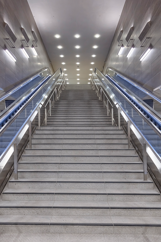 Unter den Linden station escalator corridor showing three-level vertical circulation from 14-meter-deep U5 platform to surface where U6 line crosses above, WE-EF DAC240 LED surface-mounted luminaires with integrated electronic control gear mounted on lift systems for maintenance access above escalators, identical appearance and 3000K warm white lighting technology creating uniform calm restful atmosphere designed by High Light Berlin lighting planner Thomas Spreen