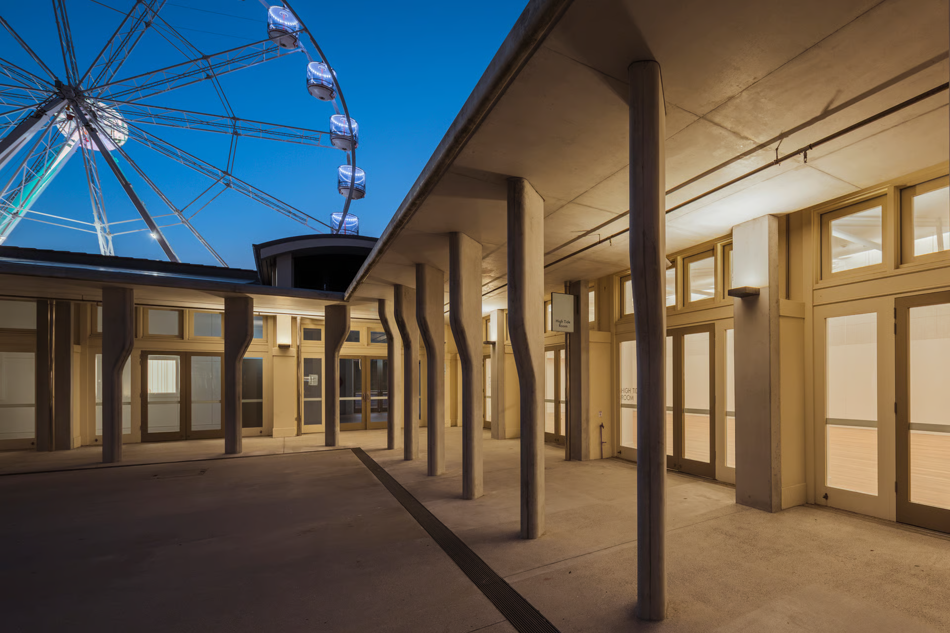 Bondi Pavilion covered colonnade at blue hour showing rhythmic sculptural timber columns, illuminated Ferris wheel visible beyond, WE-EF PLS400 wall luminaires mounted under eaves in upward orientation providing soft ceiling illumination shielded from view, demonstrating multifaceted lighting strategy balancing heritage preservation with modern community hub functionality