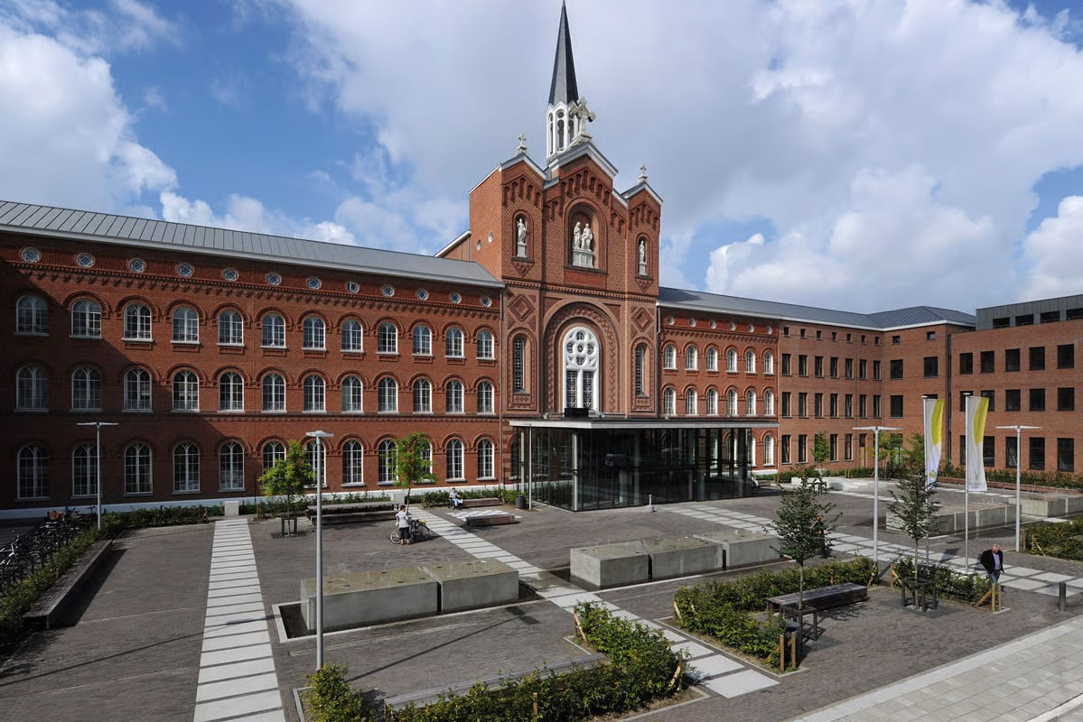 Wide-angle view of the forecourt plaza at St. Vincentius Hospital Antwerp, showing VFL540 LED pole luminaires, paved surfaces, planted green zones, and the full width of the historic red brick hospital building with its central chapel tower