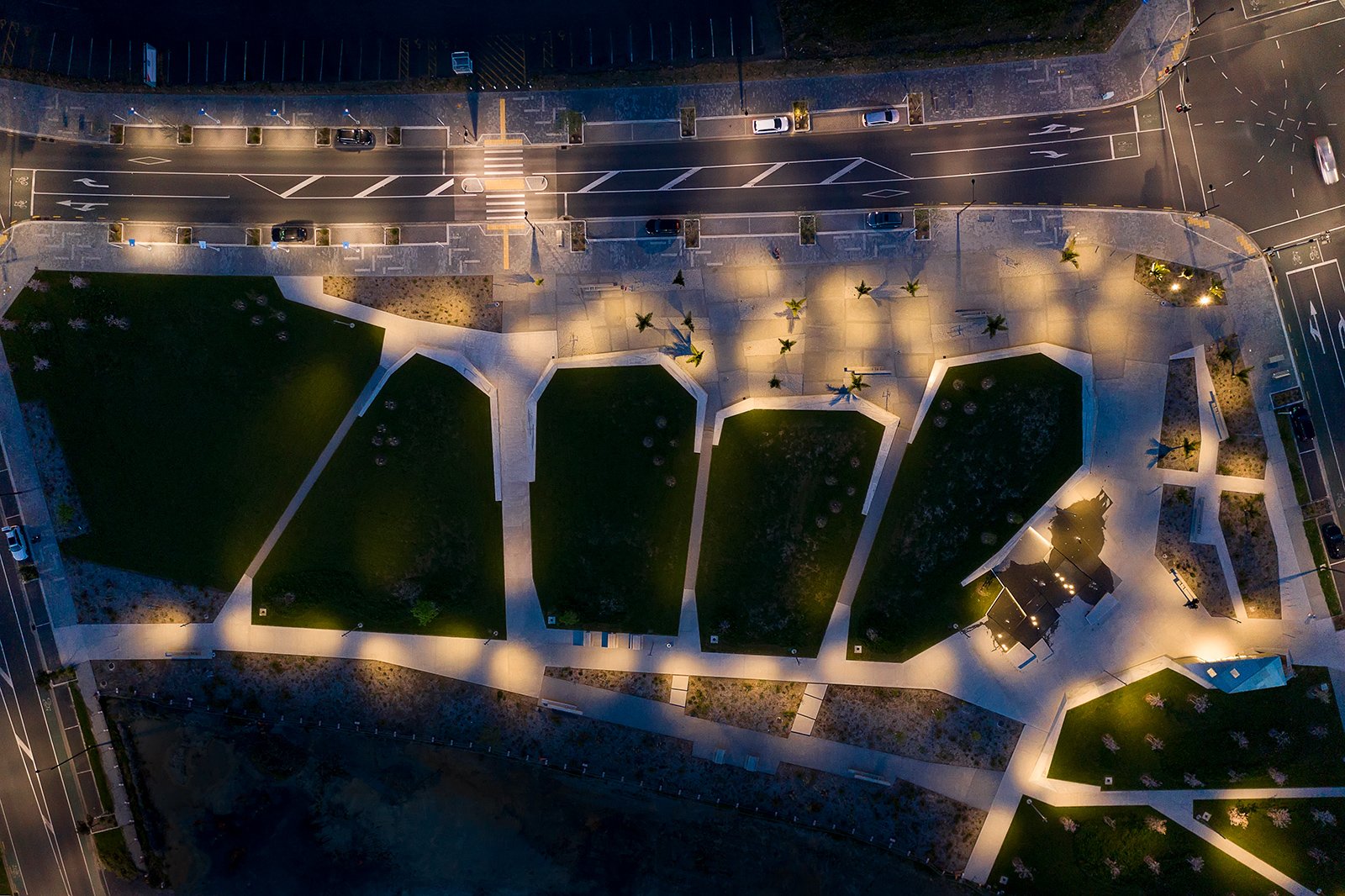 Aerial night view of Te Hauāuru Park Westgate showing organic lawn patterns structured by walls inspired by upper Waitematā harbour inlets, innovative WE-EF lighting design by MHL Auckland using 66 FLC121 projectors with medium-beam optics mounted at varying heights (5.5-6.5m) on 7-meter poles creating directional pinpointed illumination avoiding standard grid layout, P2 standard lighting ensuring safe thoroughfare between North West Shopping Centre (one of Australasia's largest precincts) and apartments in rapidly developing Auckland town centre 20km west of city