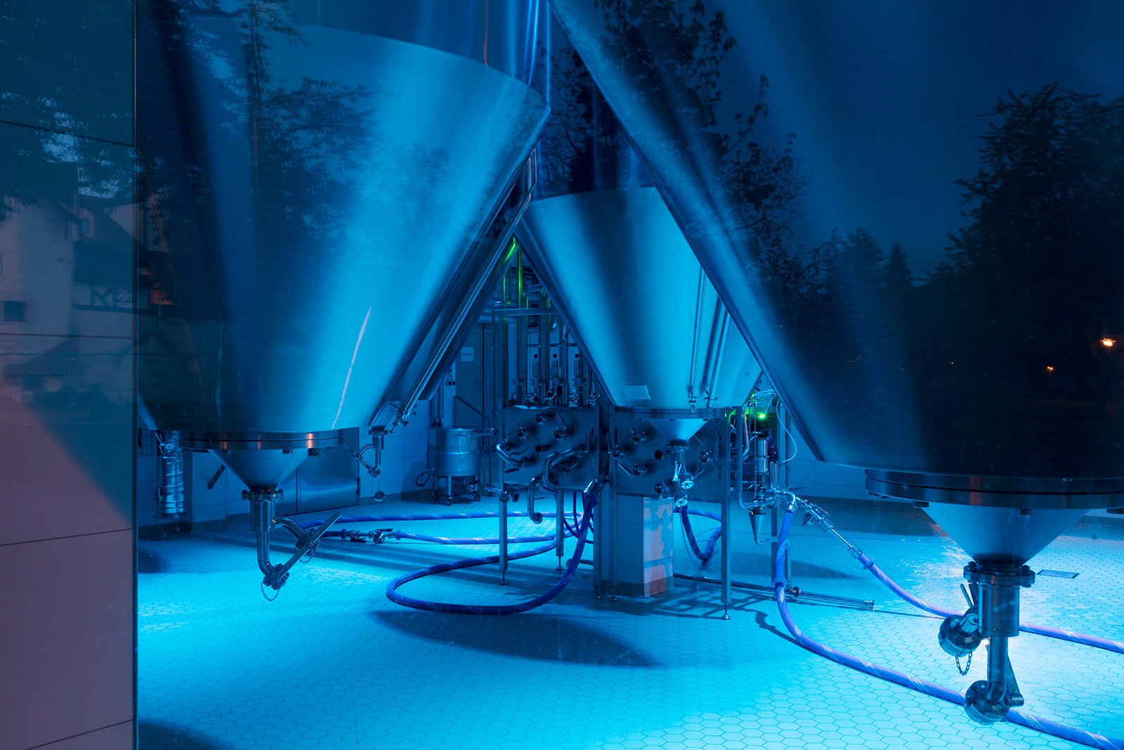 Schattenhofer Brewery fermentation tank room illuminated in atmospheric cyan-blue using WE-EF FLC220-CC RGBW projectors with IP66 splashproof rating, stainless-steel brewing equipment visible through large panoramic windows creating dramatic nighttime spectacle with DMX-controlled automatic color changes, showcasing how 300-year-old traditional Bavarian family brewery in Beilngries embraces futuristic lighting design to signal transparency, quality and innovation while competing in modern craft beer market