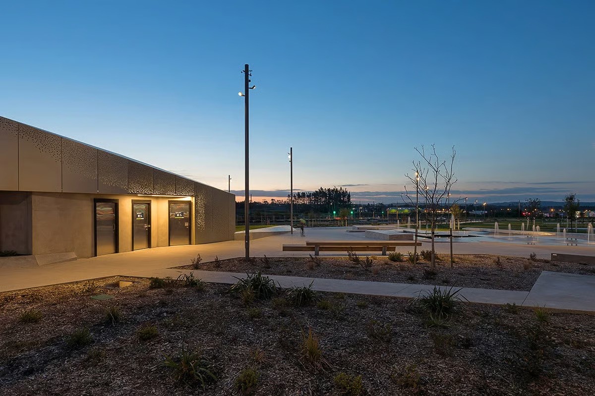 Te Hauāuru Park small amenity building at twilight in Westgate town centre 20 kilometers west of Auckland, WE-EF lighting providing safe thoroughfare illumination for 1.1-hectare green heart connecting massive North West Shopping Centre with apartments and office buildings, design by Isthmus landscape architects taking cues from upper Waitematā harbour inlets and historic kauri forests, comprehensive lighting scheme by MHL Auckland using 66 WE-EF FLC121 12W projectors on 7-meter poles avoiding grid pattern