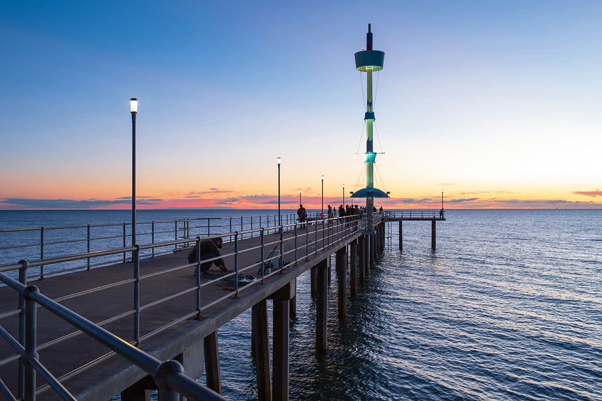 Brighton Jetty extending into Gulf St Vincent at sunset, showing the complete lighting installation with WE-EF ZFT434 luminaires mounted on poles along the wooden pier, and visitors gathered near the distinctive green observation tower at the jetty's end