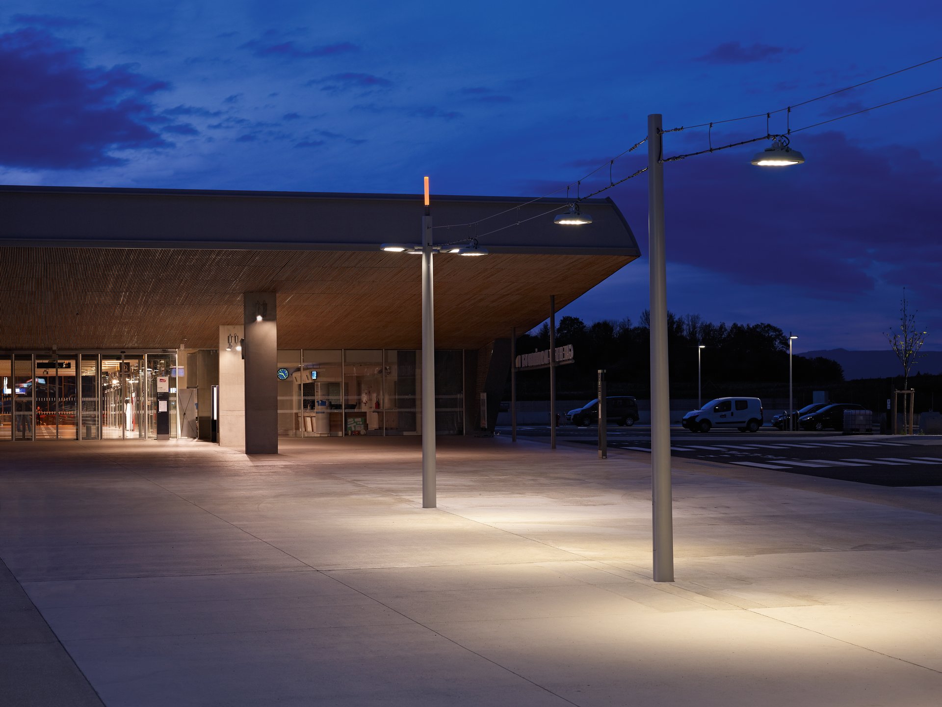 Night view of the entrance area of Belfort-Montbéliard TGV station with WE-EF RFL LED catenary luminaires and pole-mounted lights warmly illuminating the forecourt with its timber soffit