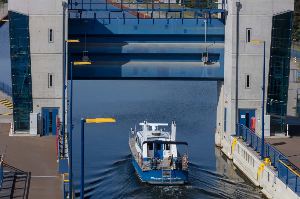 Wasserschutzpolizei (water police) vessel entering Niederfinow ship lift trough during daytime operation, showing 12.50-meter-wide by 4-meter-deep trough entry with blue holding gates, exposed concrete pylons with glass elevator shafts, golden yellow safety fenders and accents complementing blue and gray color scheme, WE-EF PFL540 luminaires visible on poles designed for optimal wall illumination critical to safe vessel entry and egress on Oder-Havel waterway infrastructure