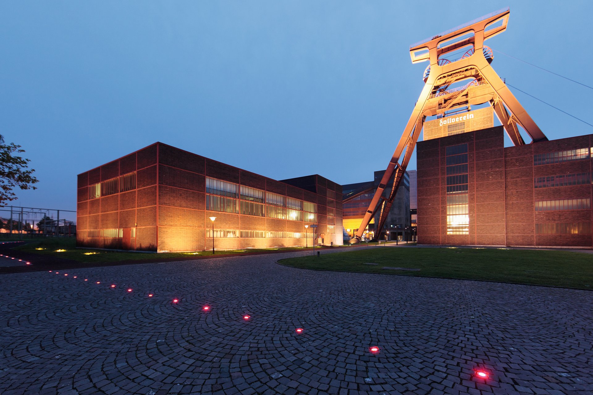 Dusk view of Zeche Zollverein in Essen with the warmly illuminated headframe and brick buildings, and a curved row of red ETC119 inground uplights set into the cobblestone forecourt