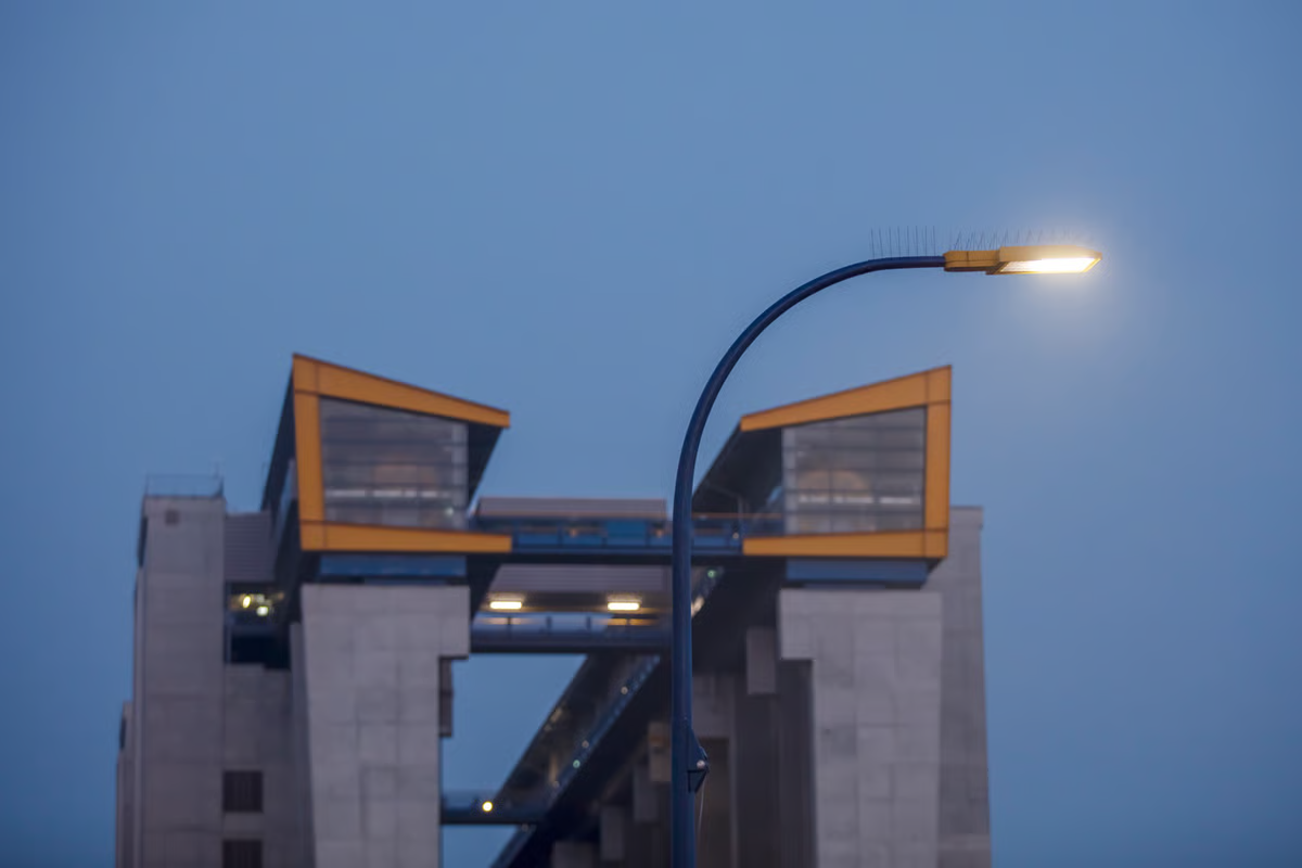 Close-up of WE-EF PFL540 LED street luminaire with custom golden yellow powder-coat finish (RAL 1004) mounted on curved blue tubular steel pole at Niederfinow ship lift, control station with yellow-framed windows visible 50 meters above on eastern pylons of exposed concrete structure, demonstrating color scheme designed by architect Udo Beuke to accent monumental engineering infrastructure at twilight