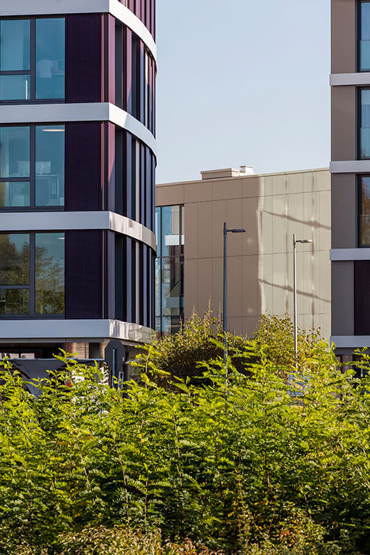 Architectural detail of Q.West Ingolstadt buildings featuring curved facades with vertical striped cladding, white balcony accents, and WE-EF pole-mounted luminaires visible against landscaped greenery