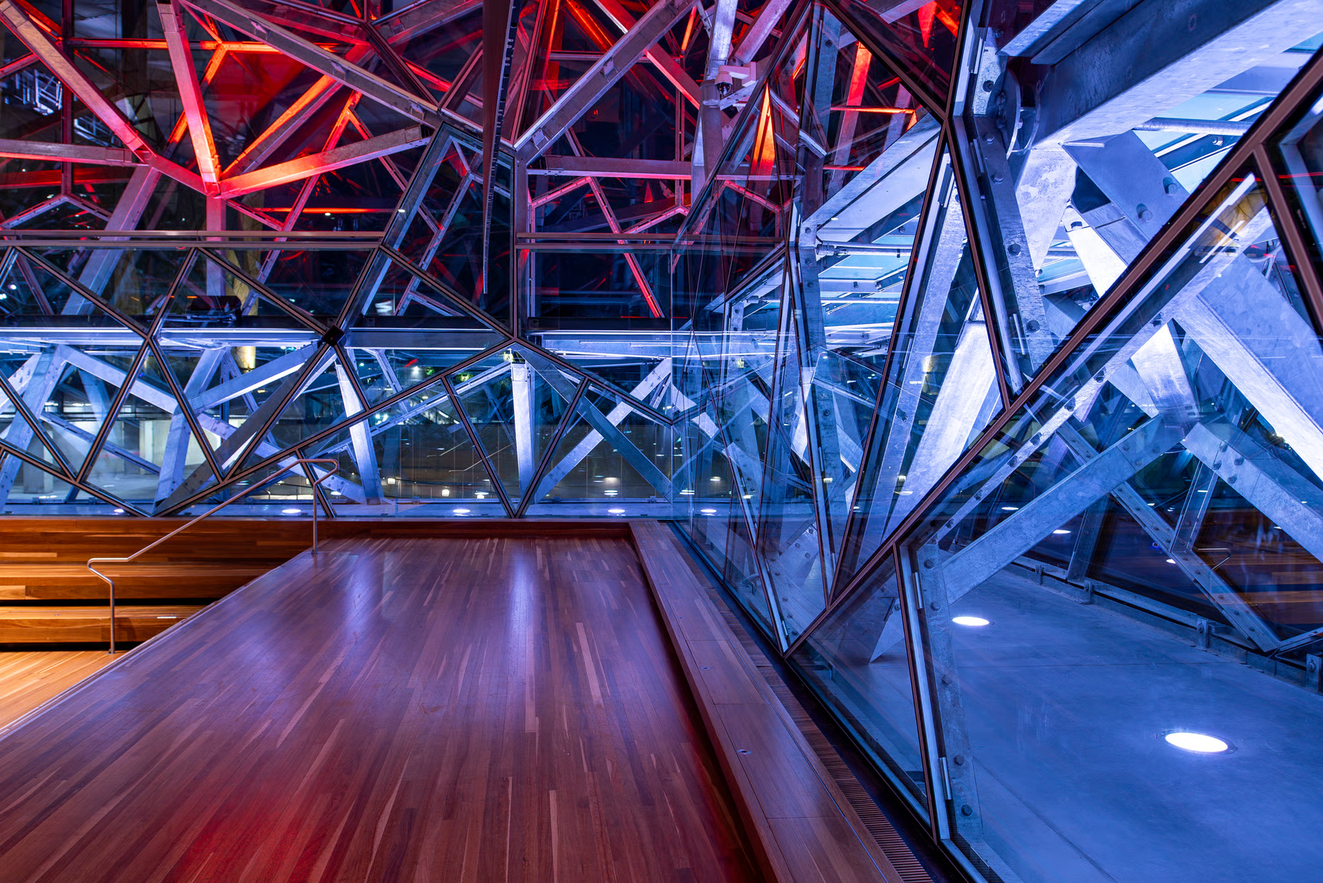 Federation Square interior architectural detail showing complex triangulated steel truss framework illuminated with WE-EF RGBW color-changing projectors creating dramatic red and blue color washes, warm wooden flooring and tiered seating visible below, glass balustrade revealing multiple structural layers and the iconic geometric construction of Melbourne's cultural landmark