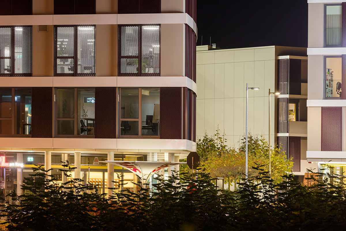 Close-up of Q.West Ingolstadt building entrance at night showcasing WE-EF ETC330-GB inground luminaires illuminating covered walkway and columns, with warm lighting creating inviting atmosphere for outdoor dining areas