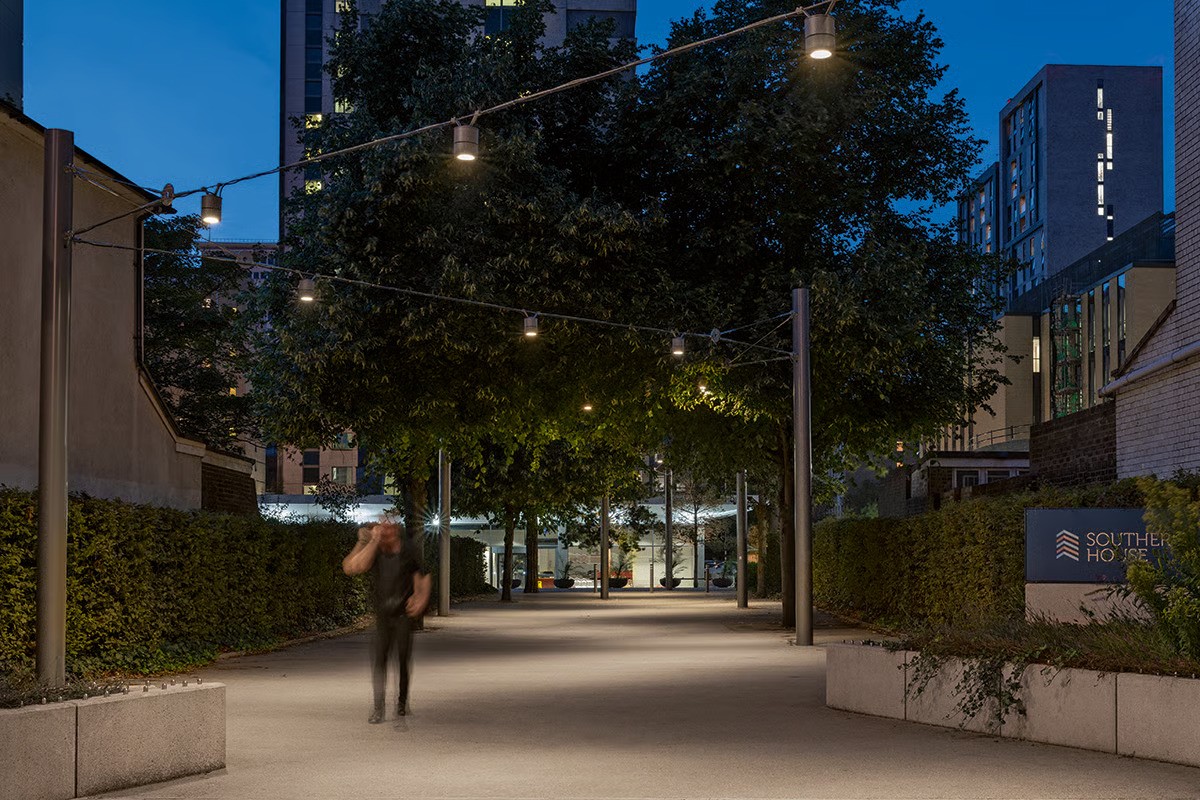 Southern House Croydon entrance pathway at night with motion-blurred pedestrian showing WE-EF DAC220 LED catenary lighting on zigzag wire cable pattern intuitively guiding visitors from George Street to 1960s office complex, warm 3000K illumination creating lively pleasant atmosphere with taut cable system engineered by local WE-EF office, landscape design by LUC Land Use Consultants for LCR Property 2018 rejuvenation transforming sought-after South London location with attractive pedestrian-focused infrastructure near East Croydon station