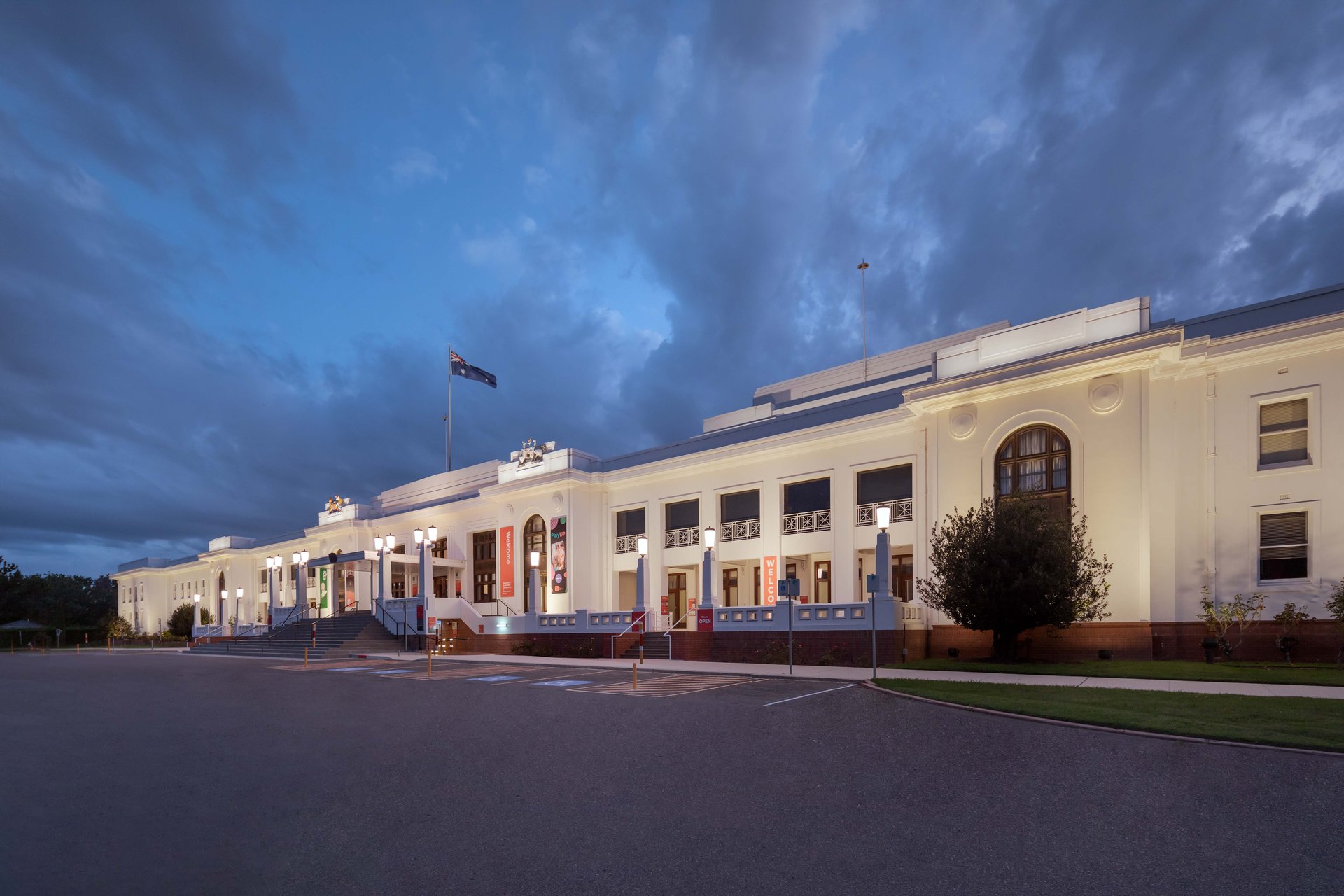 Old Parliament House at dusk showing natural white baseline illumination from WE-EF FLC200-CC and VLR100-CC LED luminaires, stripped classical architecture with characteristic arched entrance, columned portico, and symmetrical facade designed by John Smith Murdoch as provisional Federal Parliament 1927, now National Heritage-listed Museum of Australian Democracy, dramatic storm clouds overhead, energy-efficient color-changing system designed by WSP replacing inefficient halogen floodlights, supplied by Integral Lighting and built by Manteena contractor
