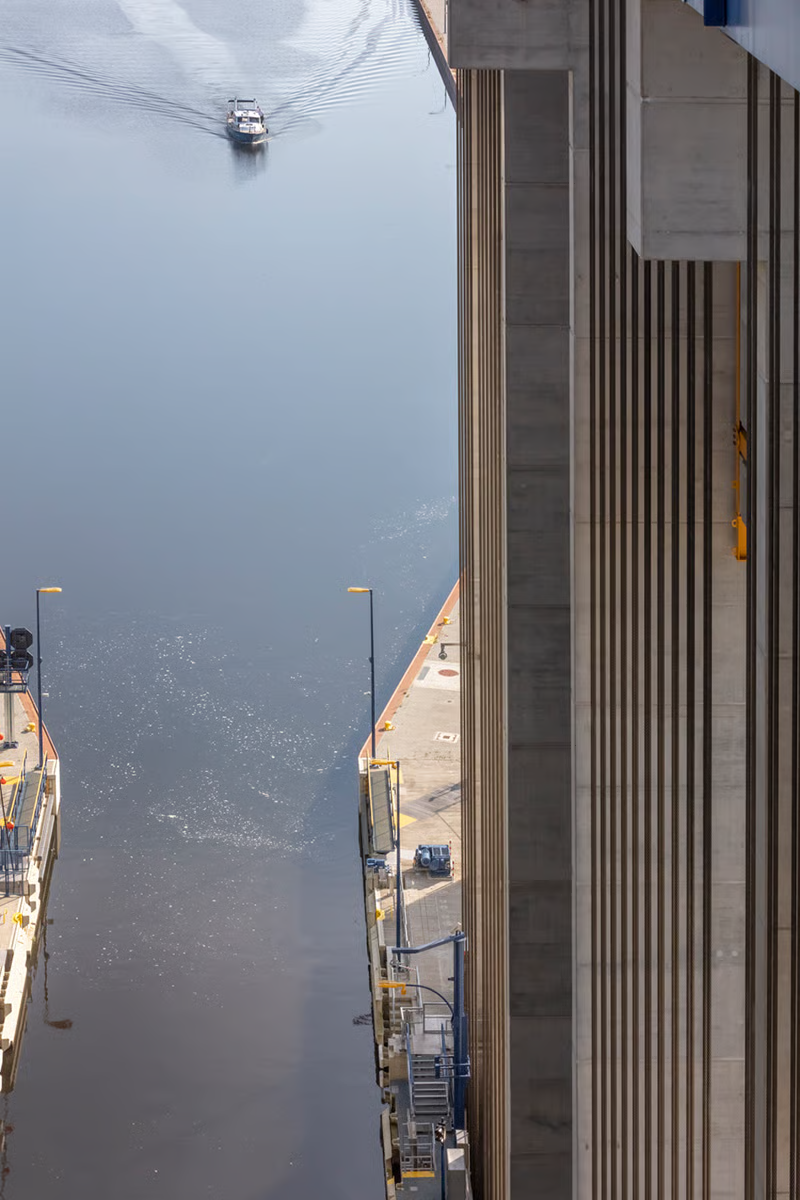 Aerial view of Niederfinow ship lift trough and lower forebay showing cargo vessel approaching entry, massive exposed concrete pylons with vertical ribbing and golden yellow safety accents, WE-EF PFL540 LED luminaires on golden yellow housings mounted on blue poles at varying heights (8m forebays, 6m trough) providing DIN 67500-compliant illumination for precision maneuvering by bargemen during 6am-10pm daily operations