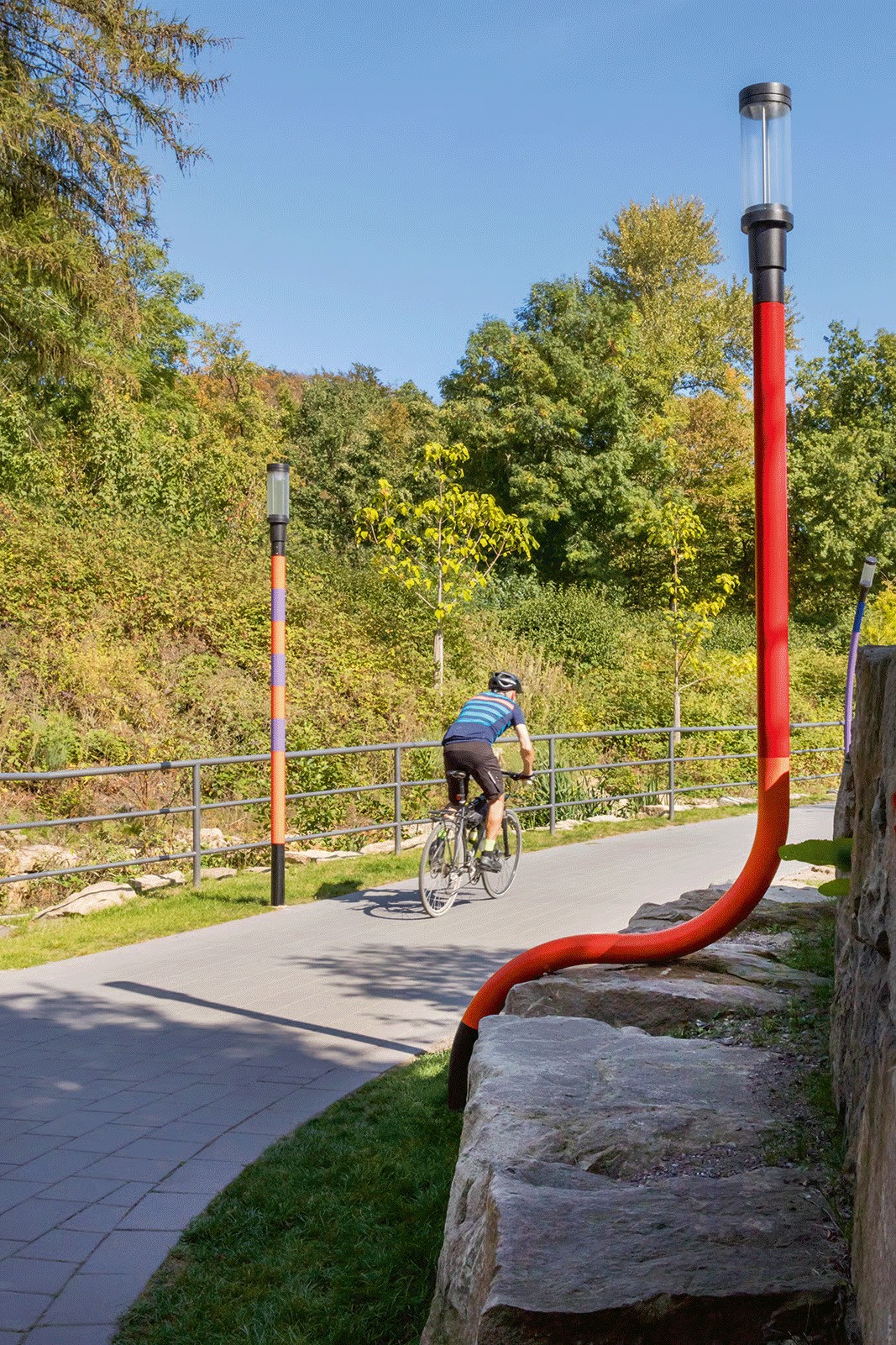 Barrier-free access ramp to Bochum railway museum tunnel showing dramatically curved red WE-EF ZFT440 LED luminaire seeming to walk or lean in human-like movement, cyclist riding toward Ruhr river meadows along popular cycle path connecting Bochum city center with Dahlhausen district, 2020 renovation by City of Bochum transforming formerly uneasy 2.35-meter-high passage into attractive safe thoroughfare for pedestrians and cyclists using playful light sculptures concept