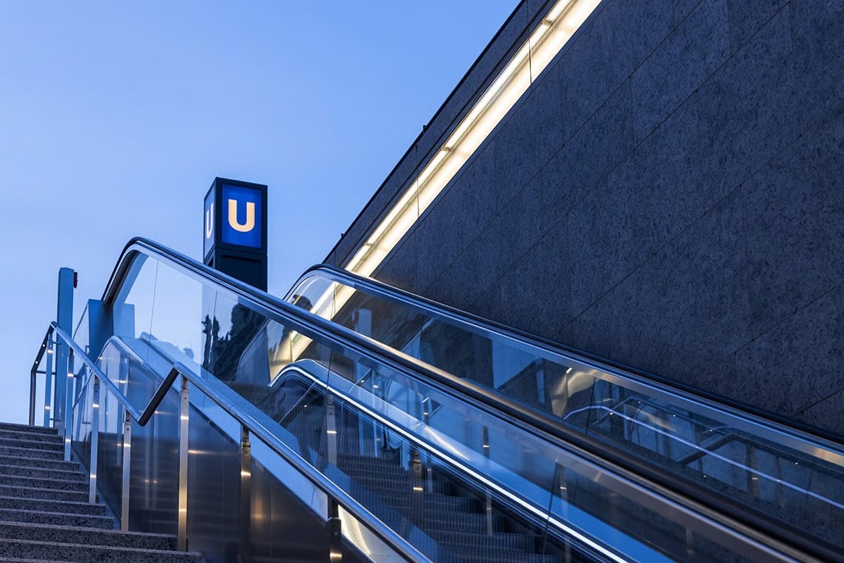 Contemporary entrance to Museumsinsel U5 station featuring minimalist granite parapet design with integrated WE-EF VLR100 series linear luminaires providing functional downward wall wash lighting into entrance void, transparent glass-enclosed escalator descent visible beneath iconic Berlin U-Bahn signage, architectural lighting solution by Thomas Spreen/High Light supporting Max Dudler's design concept for 2020-completed station serving Museum Island's cultural heart beneath Spree Canal